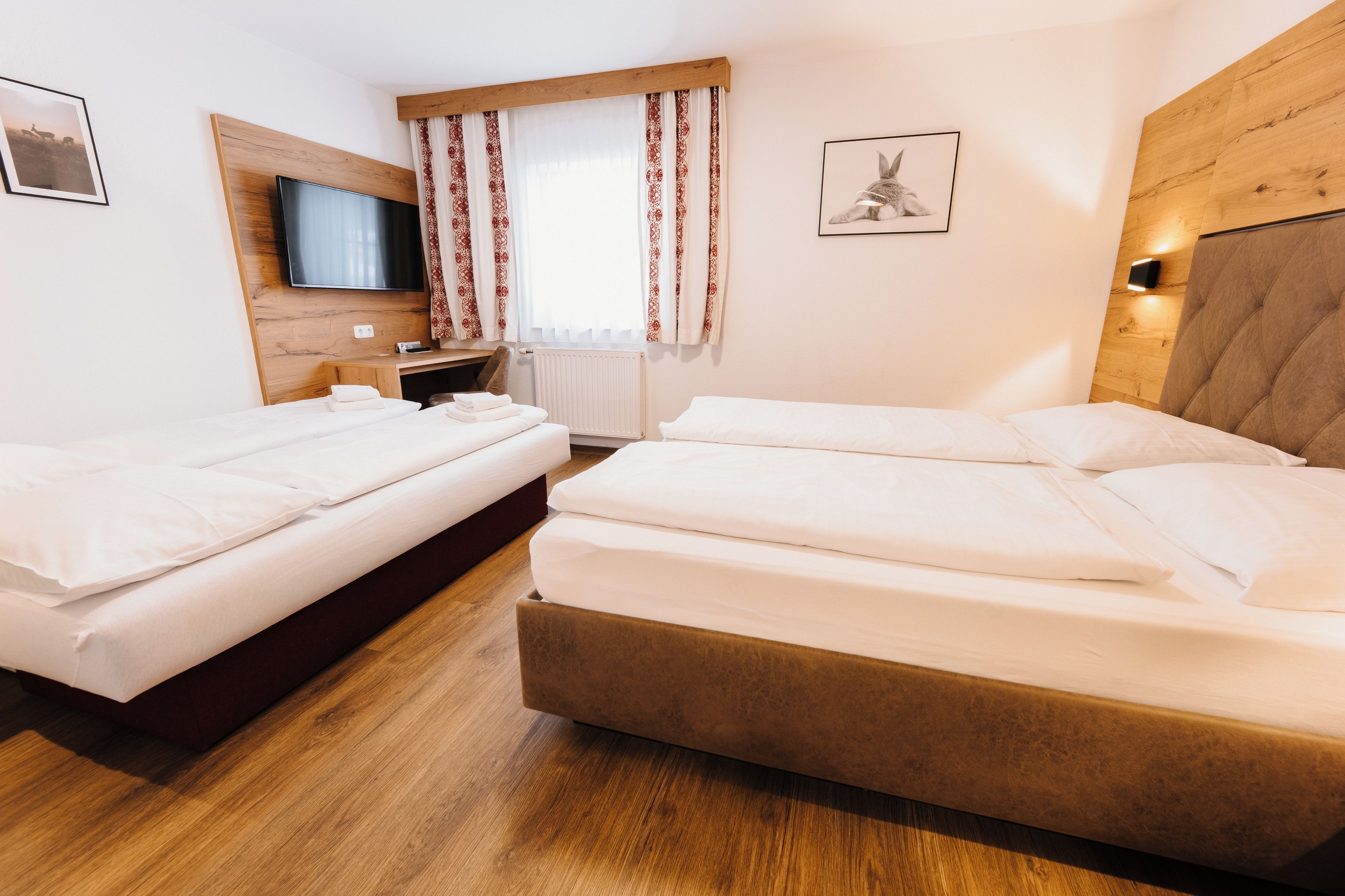 Quarto de hotel com duas camas, cabeceira de madeira, televisão e janela com cortinas estampadas.