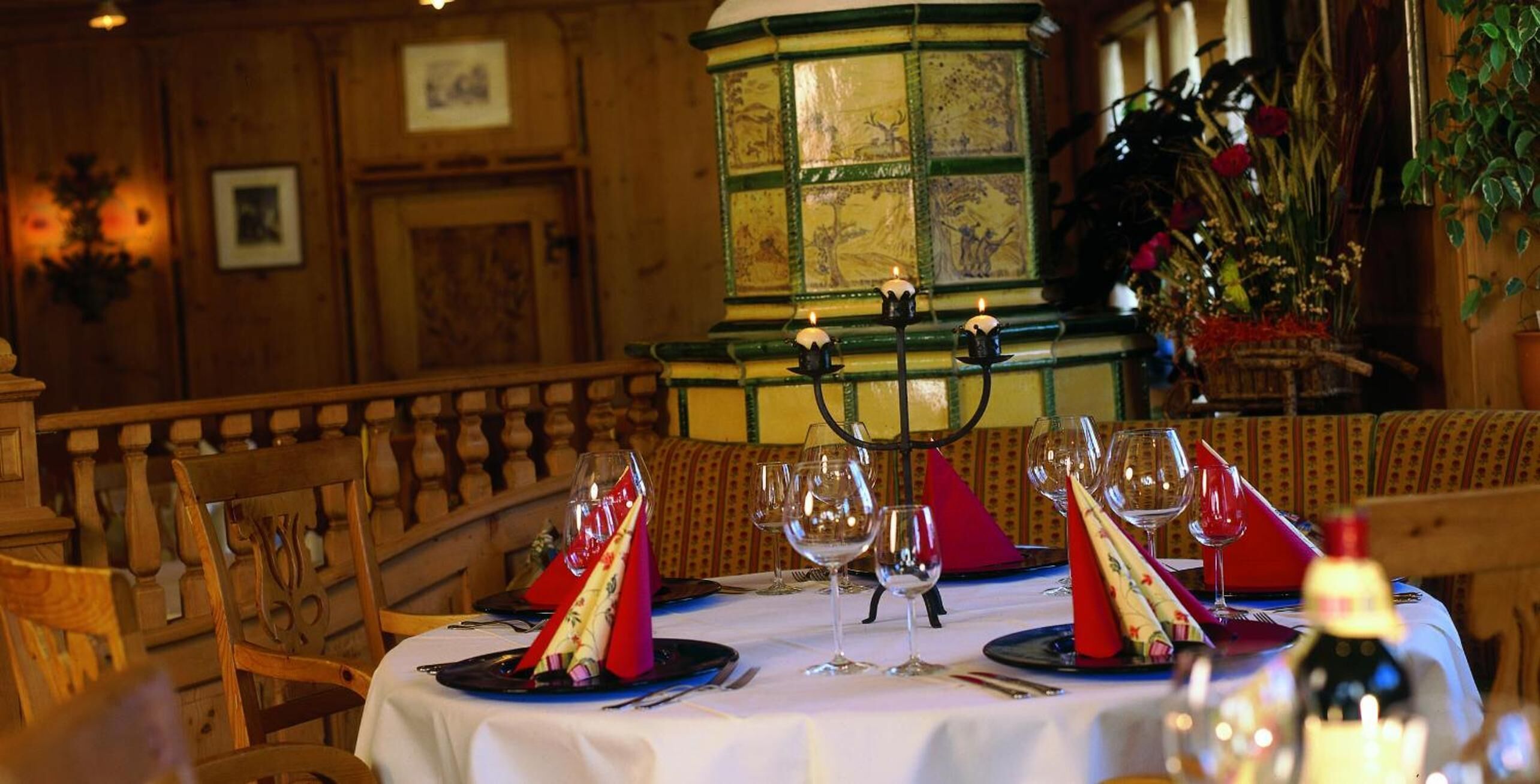 A restaurant setting with a wooden interior, a round table set for two with wine glasses and red napkins, and a candelabra with candles.
