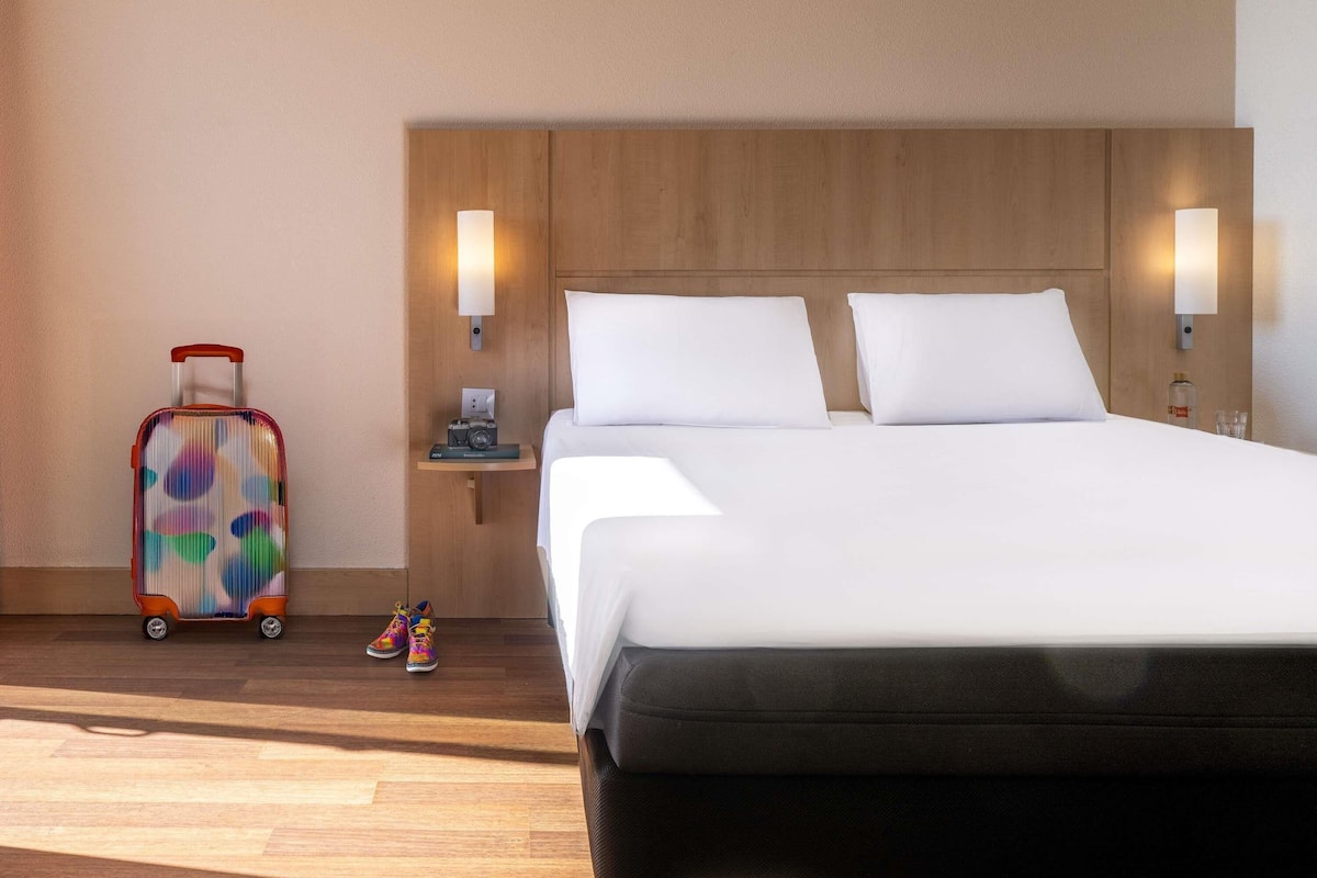 Una habitación de hotel con una cama, dos lámparas de noche, una mesita de noche con lámpara, una maleta y un par de zapatos coloridos.