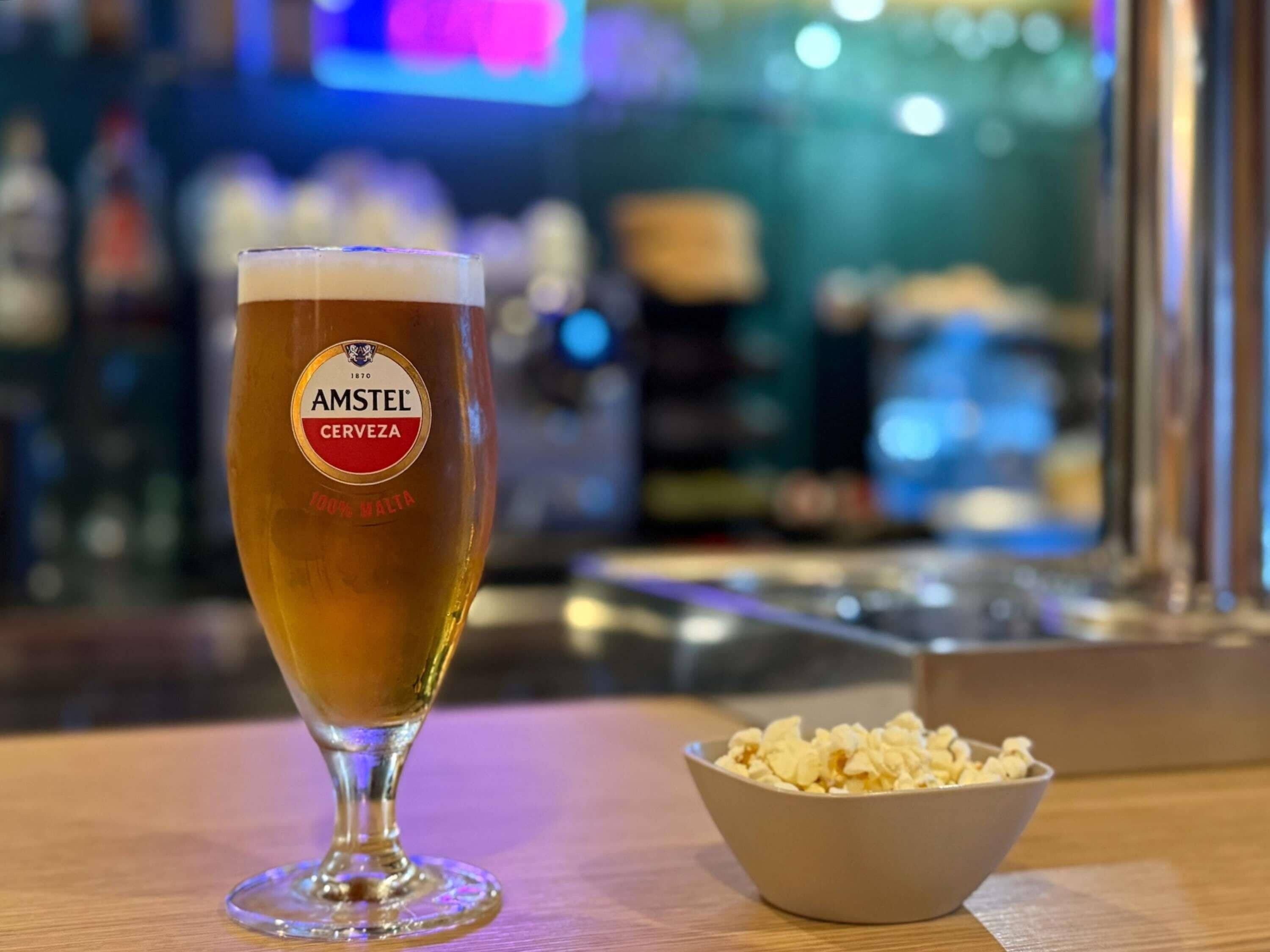 A glass of Amstel beer with a label indicating it is 100% malt, placed on a wooden table next to a bowl of popcorn.