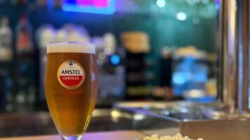 A glass of Amstel beer with a label indicating it is 100% malt, placed on a wooden table next to a bowl of popcorn.