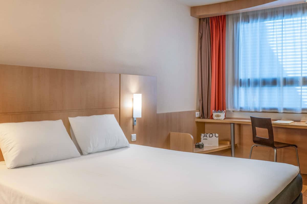 Habitación de hotel con cama, escritorio, silla, lámpara y ventana con cortinas.