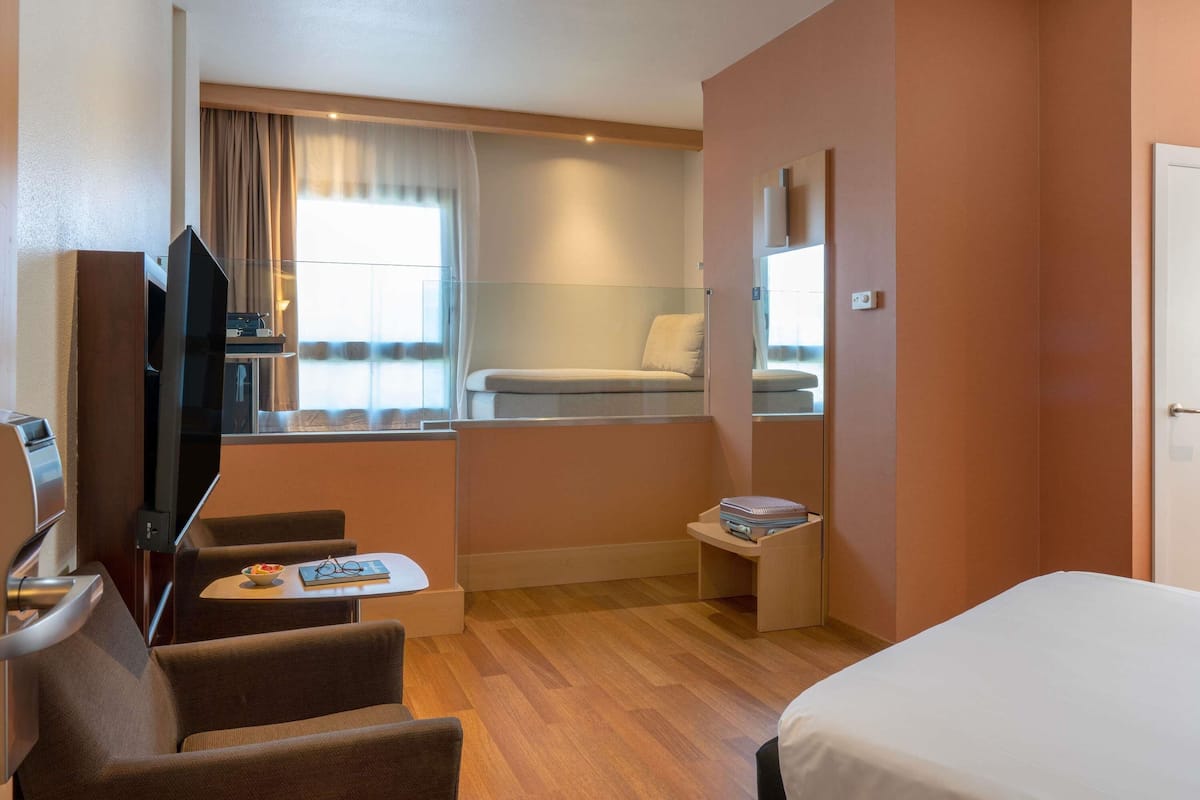 Una habitación de hotel con una cama, televisión, una pequeña mesa con un frutero y vasos, una silla, un sofá y una ventana con cortinas.