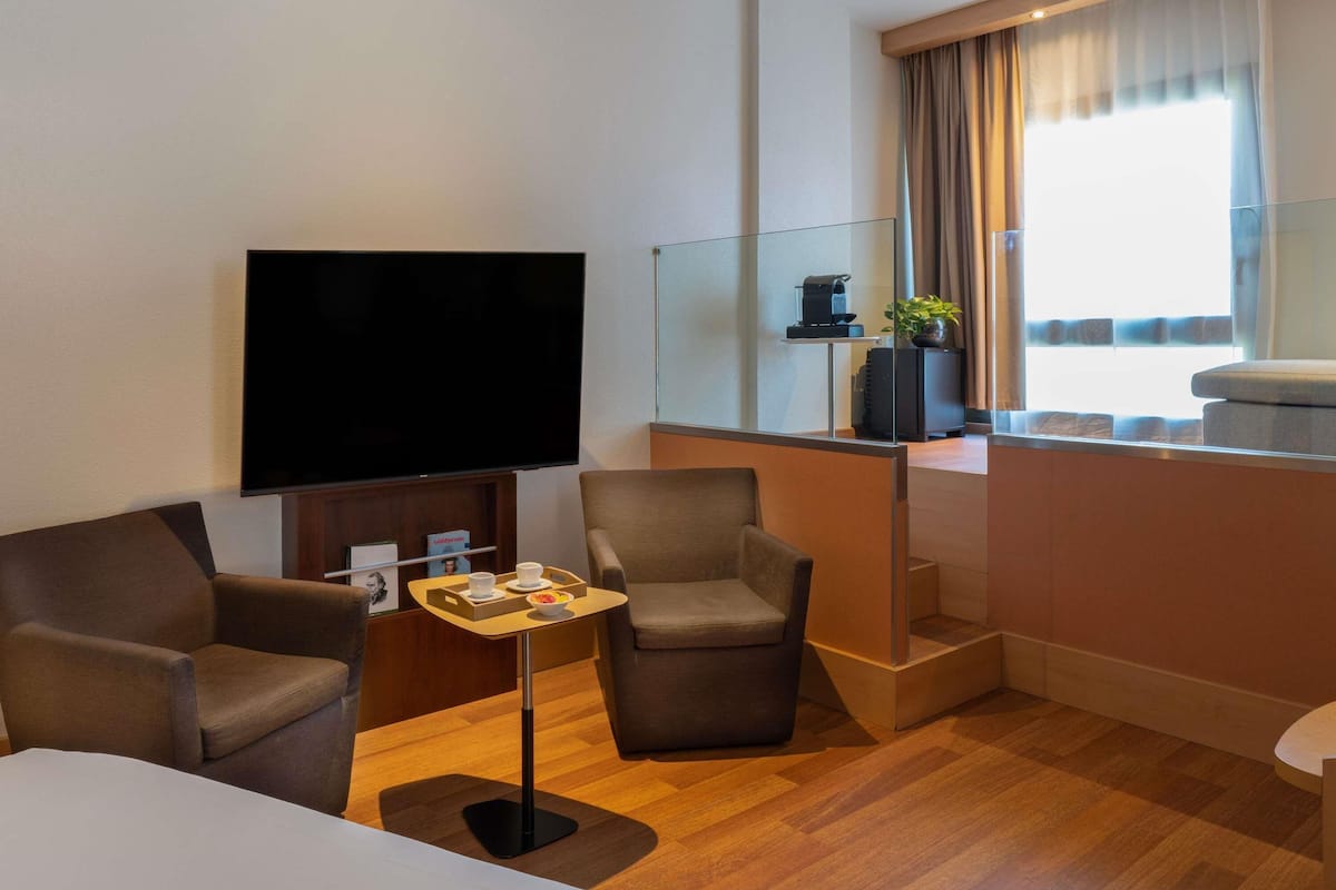 Ein Hotelzimmer mit Flachbildfernseher, zwei Sesseln, einem kleinen Tisch mit Tablett, einer Kaffeemaschine und einem Fenster mit Vorhängen.