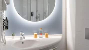 Un baño con un espejo redondo, un lavamanos, un cabezal de ducha de mano, un perchero para toallas y una pequeña planta en maceta.