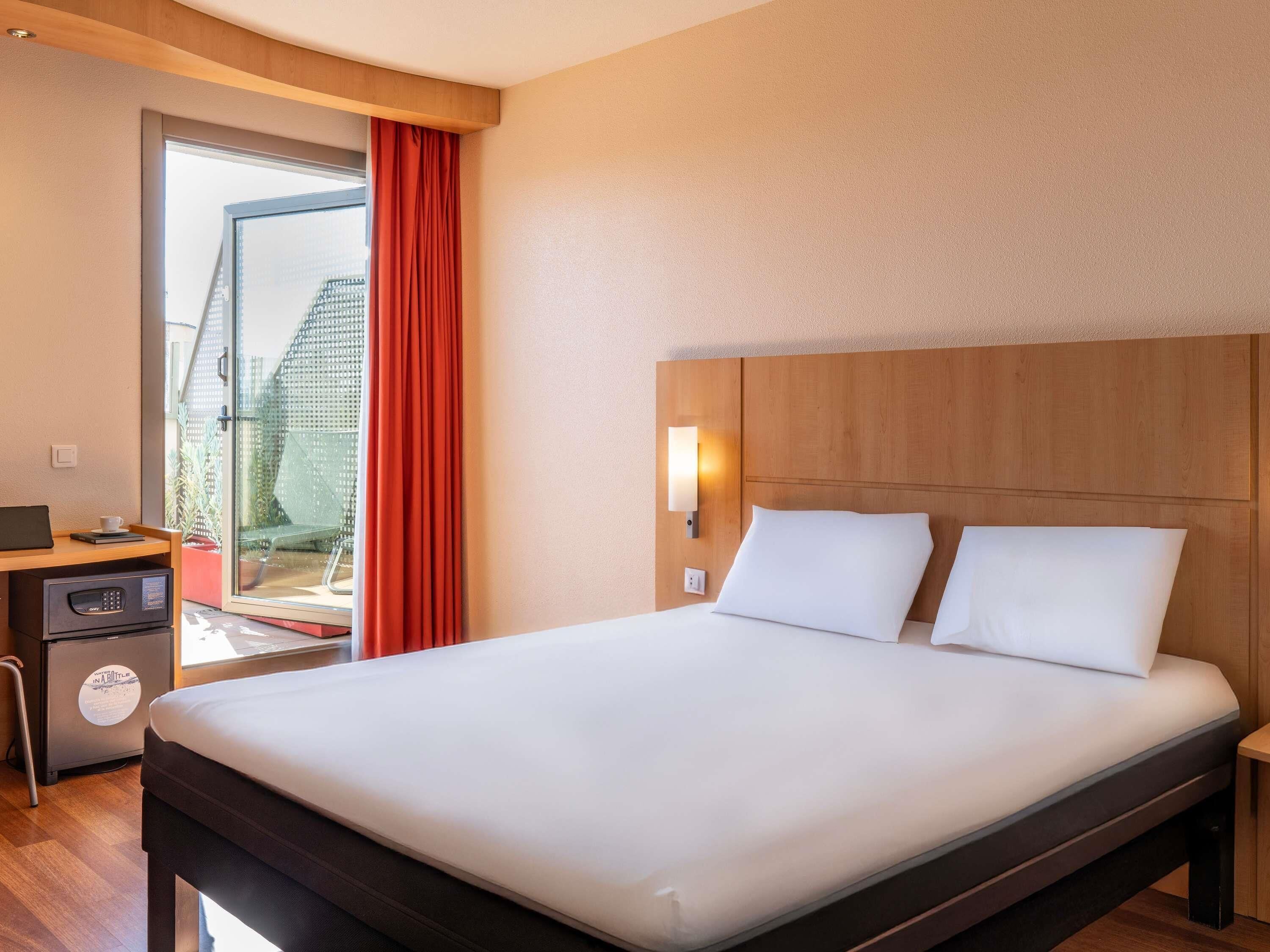 A hotel room with a bed, a desk with a computer, a small cabinet, and a window with curtains.