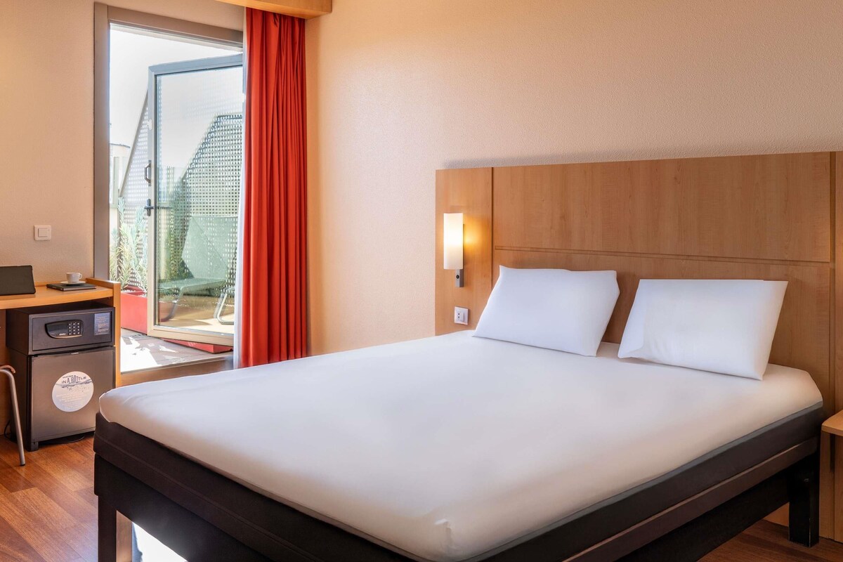 Een hotelkamer met een bed, een bureau met een computer, een klein kastje en een raam met gordijnen.