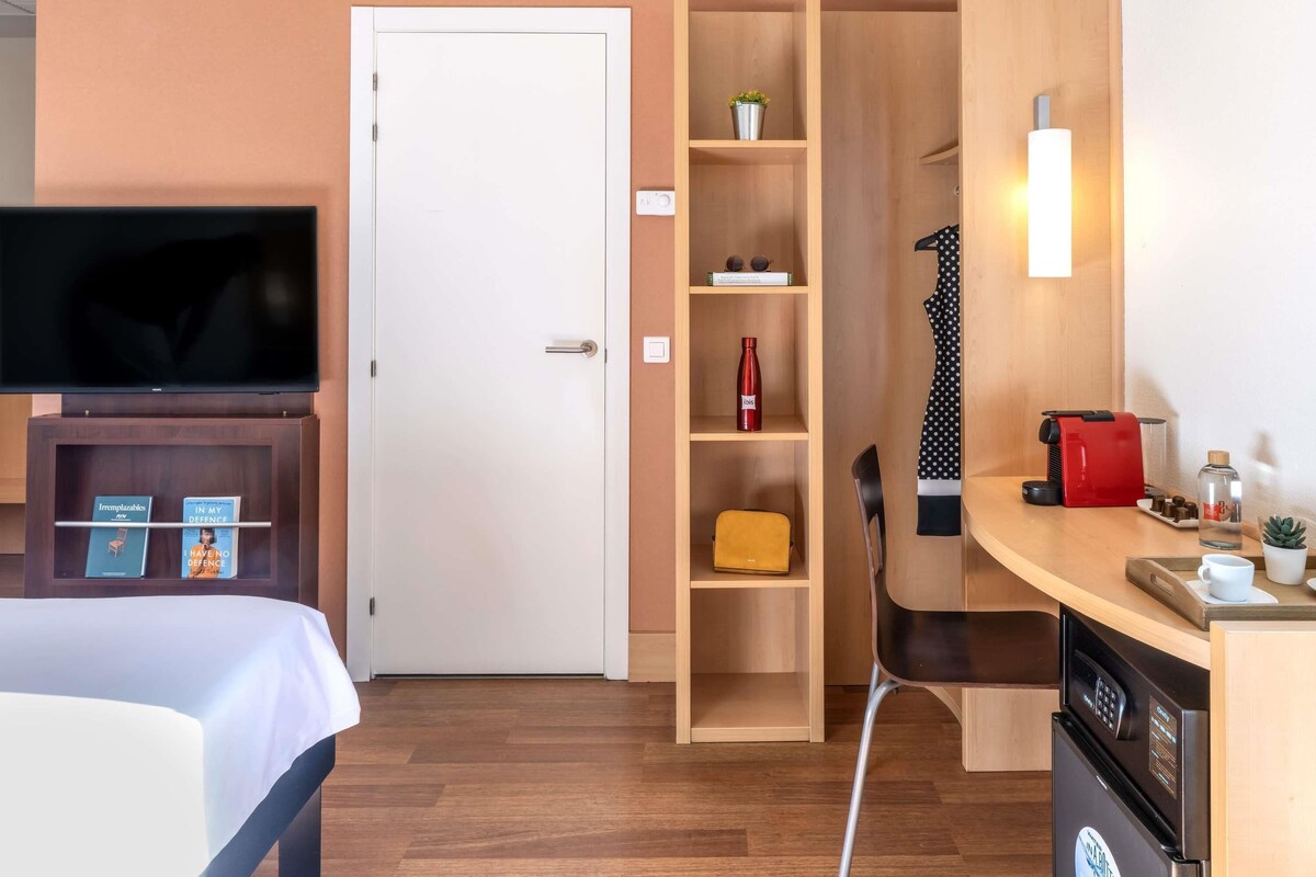 A hotel room with a bed, a TV, a wooden wardrobe, a desk with a coffee machine, and a chair.