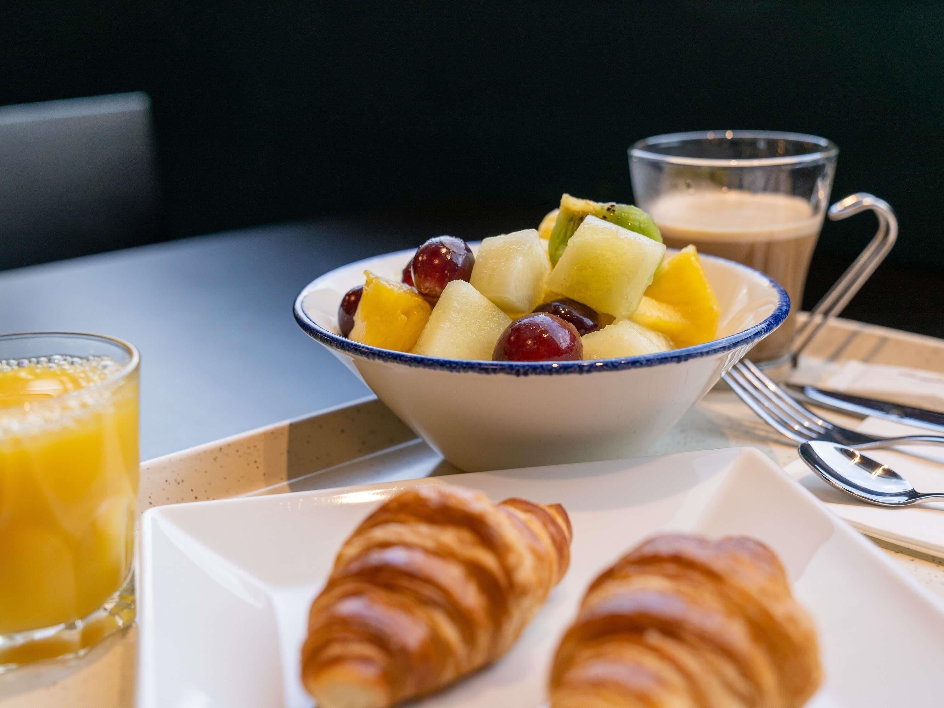 A breakfast setting with a bowl of fruit, a glass of orange juice, and two croissants on a tray.