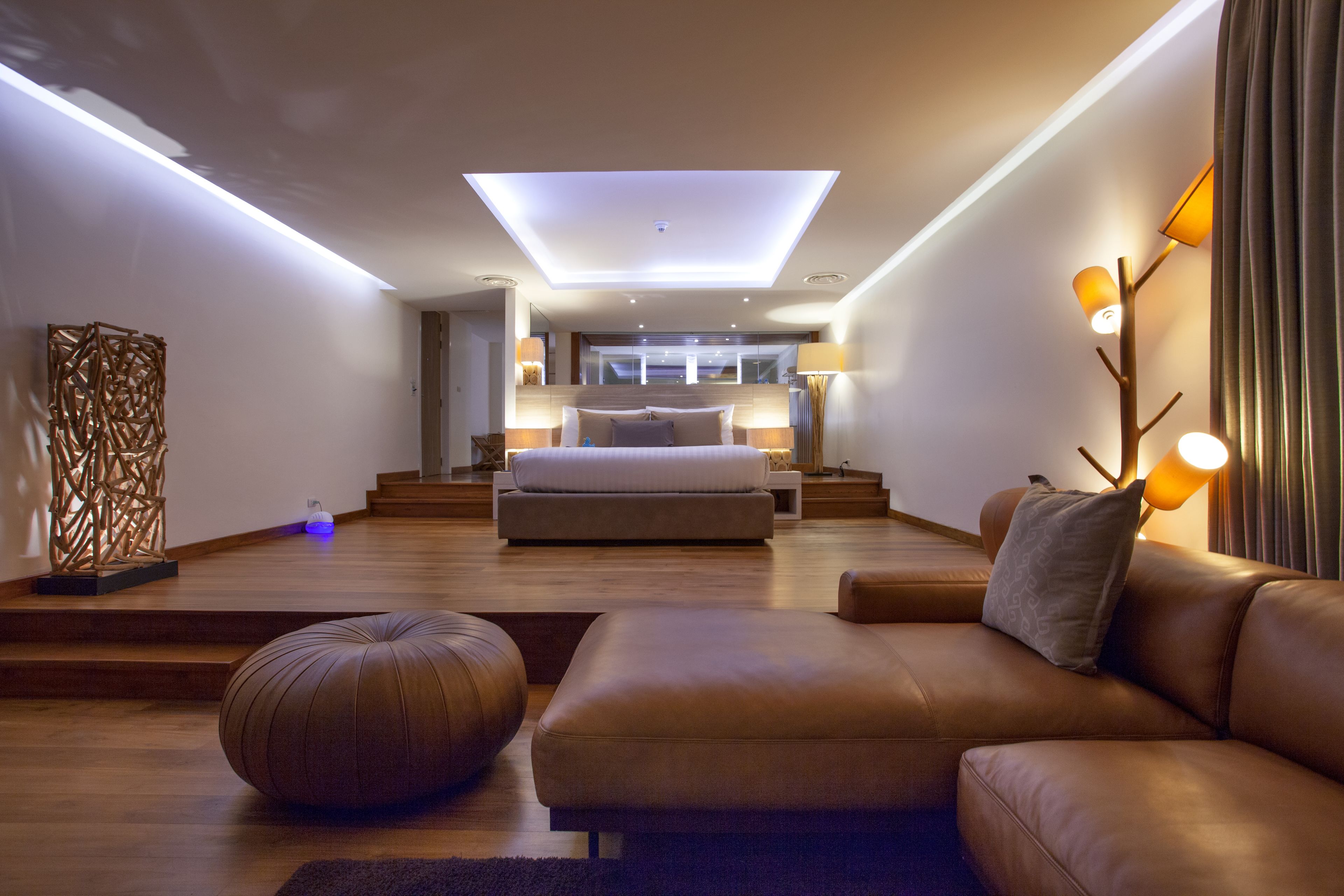 Une chambre moderne avec un grand lit, un canapé, un pouf rond et un luminaire au plafond.