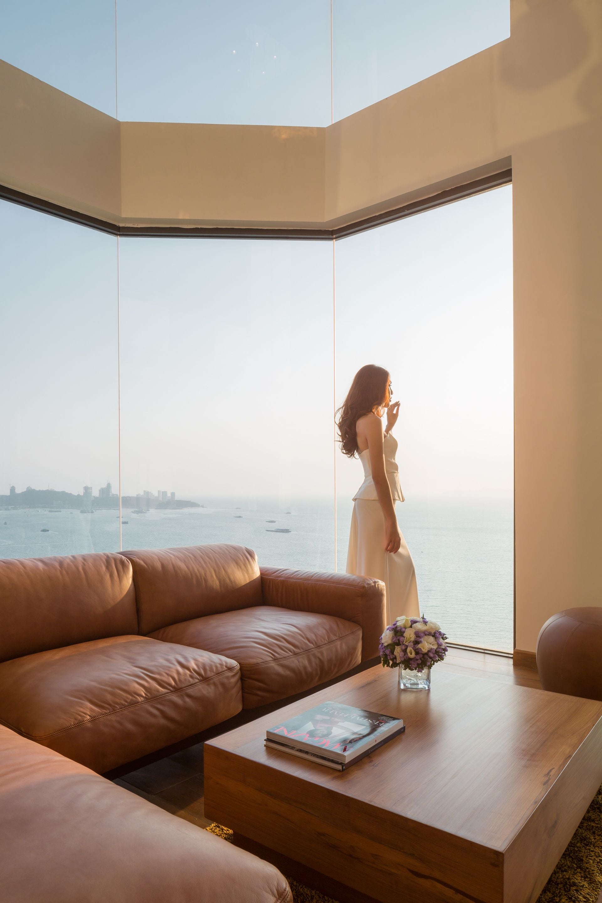 A modern living room with a large window, a leather sofa, a wooden coffee table, and a vase with flowers.