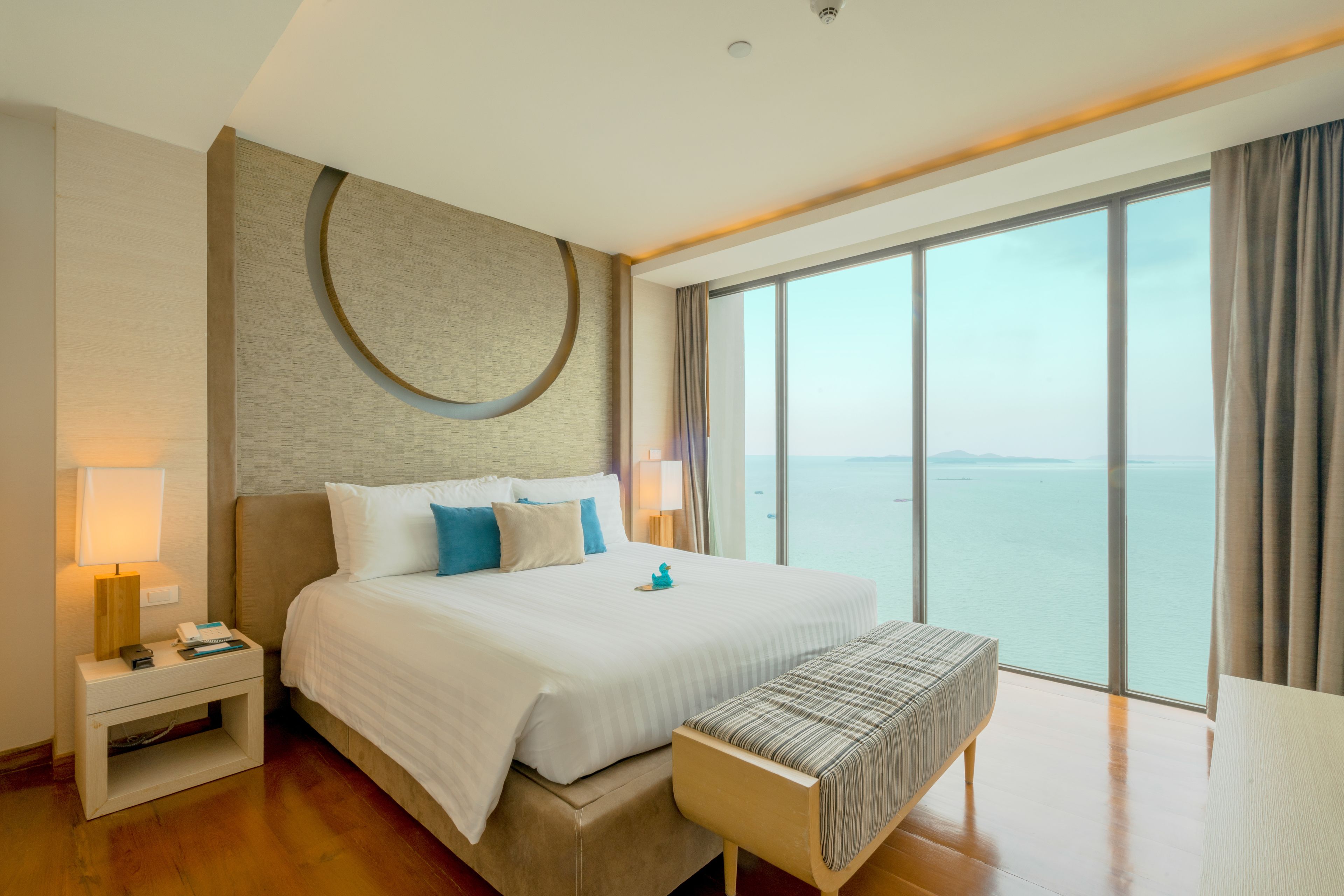 A modern hotel room with a large bed, a bedside table, a bench, and a large window offering a view of the sea.