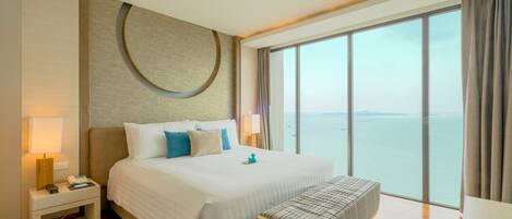 A modern hotel room with a large bed, a bedside table, a bench, and a large window offering a view of the sea.