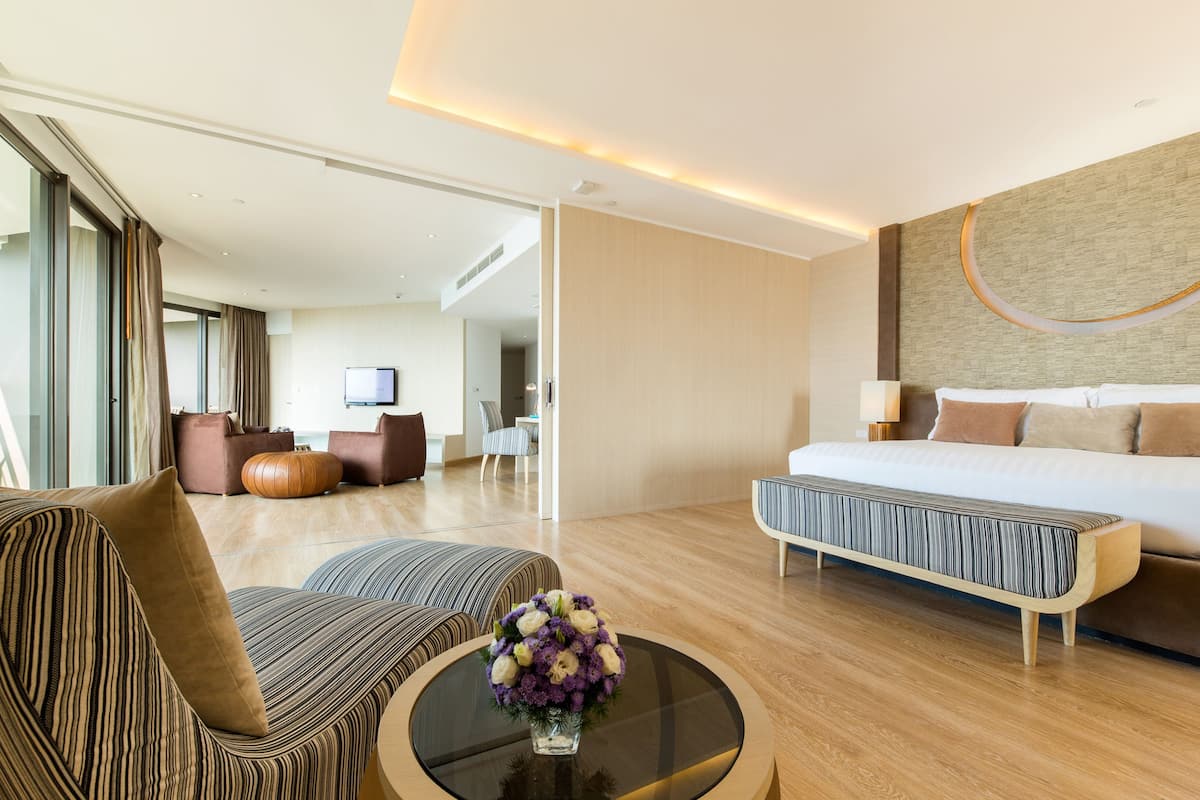 A modern hotel room with a large bed, a sofa, a coffee table with a vase of flowers, and a TV mounted on the wall.