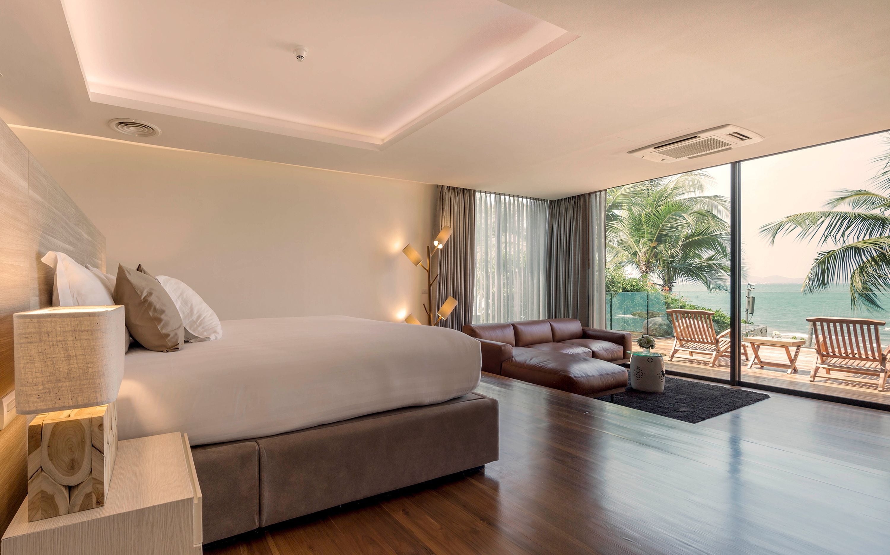 A modern hotel room with a large bed, a sofa, a wooden floor, and a balcony with a view of the sea.