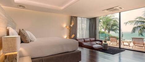 A modern hotel room with a large bed, a sofa, a wooden floor, and a balcony with a view of the sea.