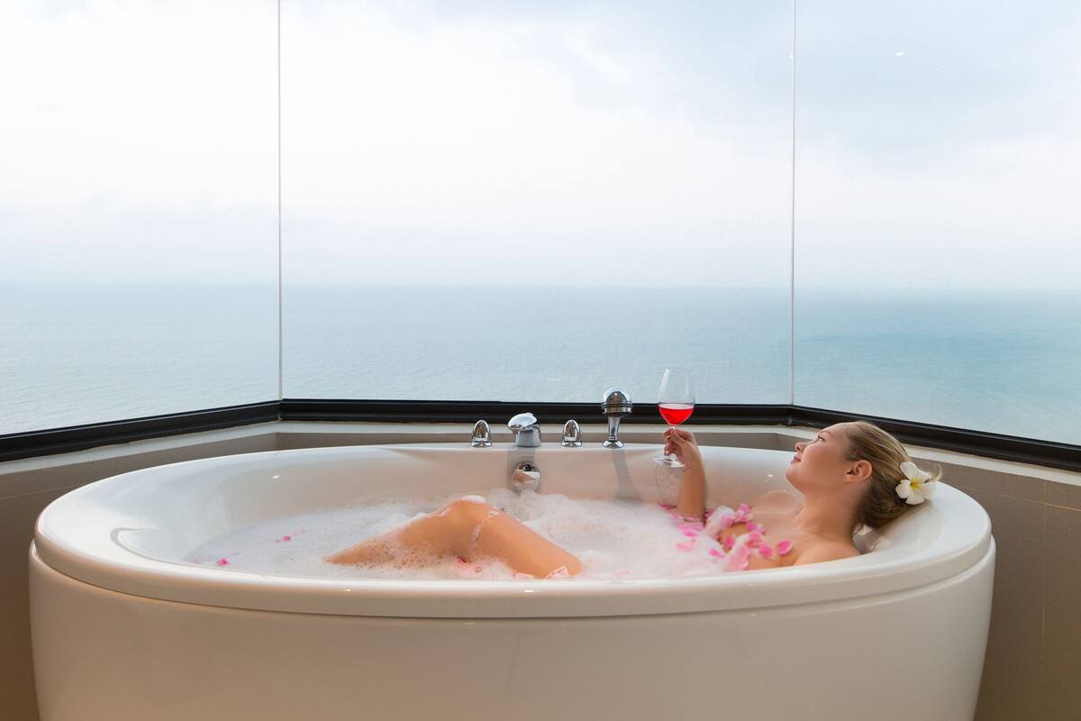 A woman relaxing in a bathtub with a glass of red wine, surrounded by bubbles and flowers, with a view of the sea.