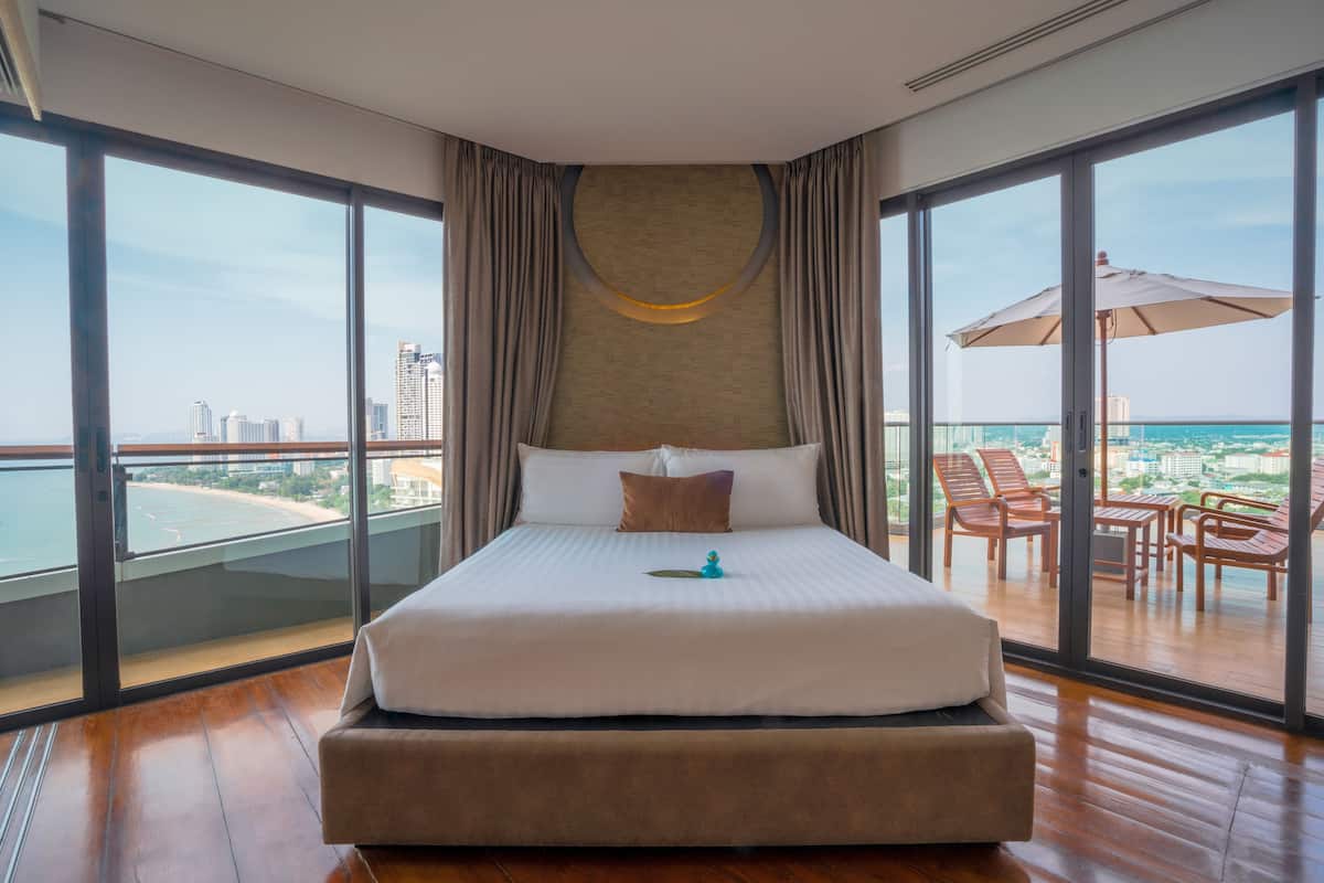 A hotel room with a large bed, a view of the city and sea, and a balcony with outdoor furniture.