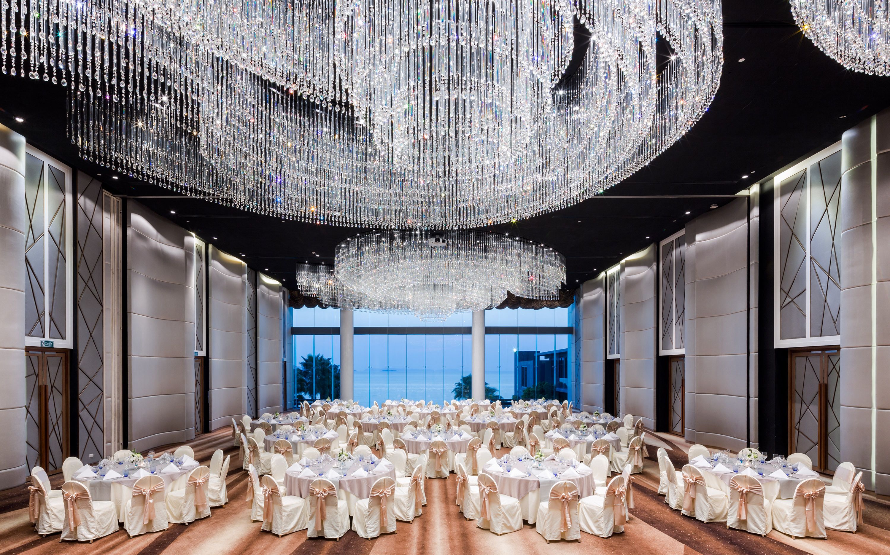 A spacious banquet hall with round tables set for a formal event, large chandeliers, and a view of the outdoors.