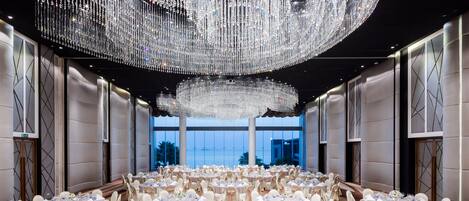 A spacious banquet hall with round tables set for a formal event, large chandeliers, and a view of the outdoors.