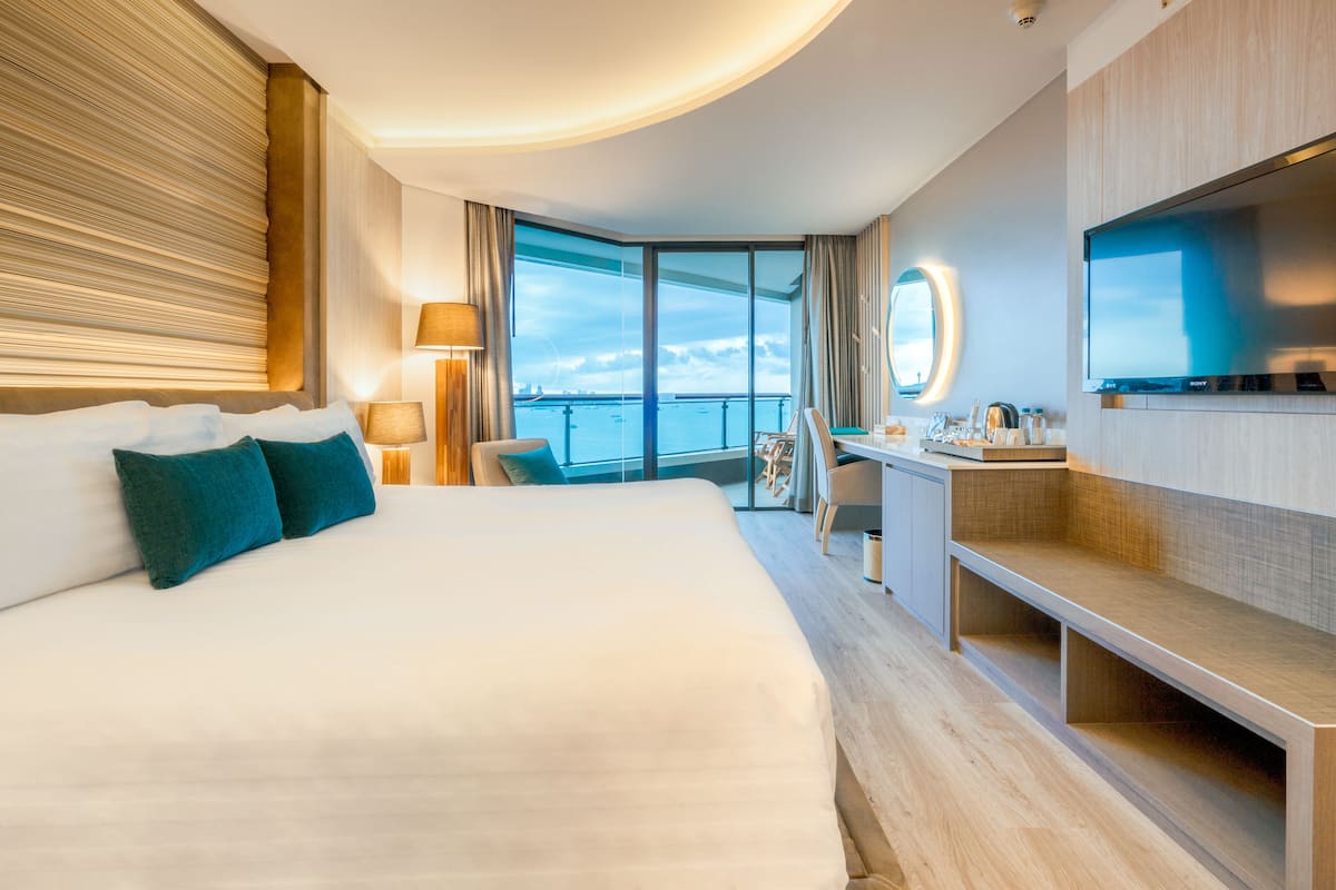 A modern hotel room with a large bed, a desk, a TV, and a balcony with a view of the sea.
