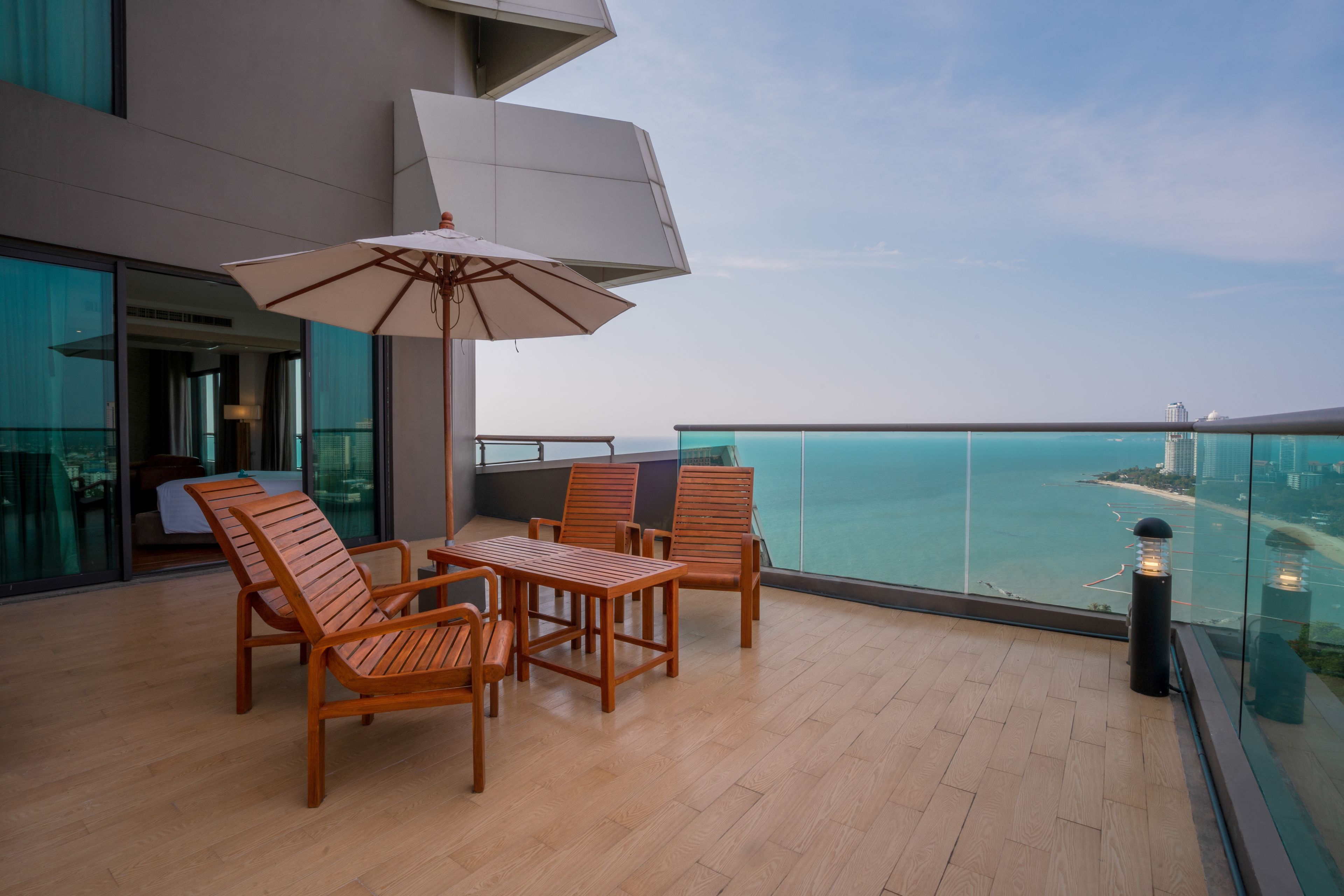 A balcony with wooden furniture, a large umbrella, and a glass railing overlooking a body of water.