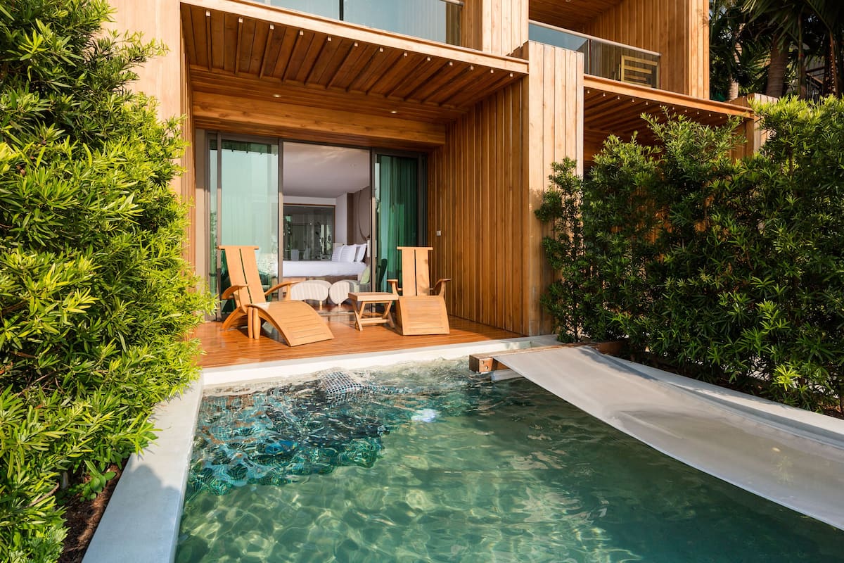 A modern outdoor pool area with wooden deck chairs and a small table, surrounded by lush greenery.