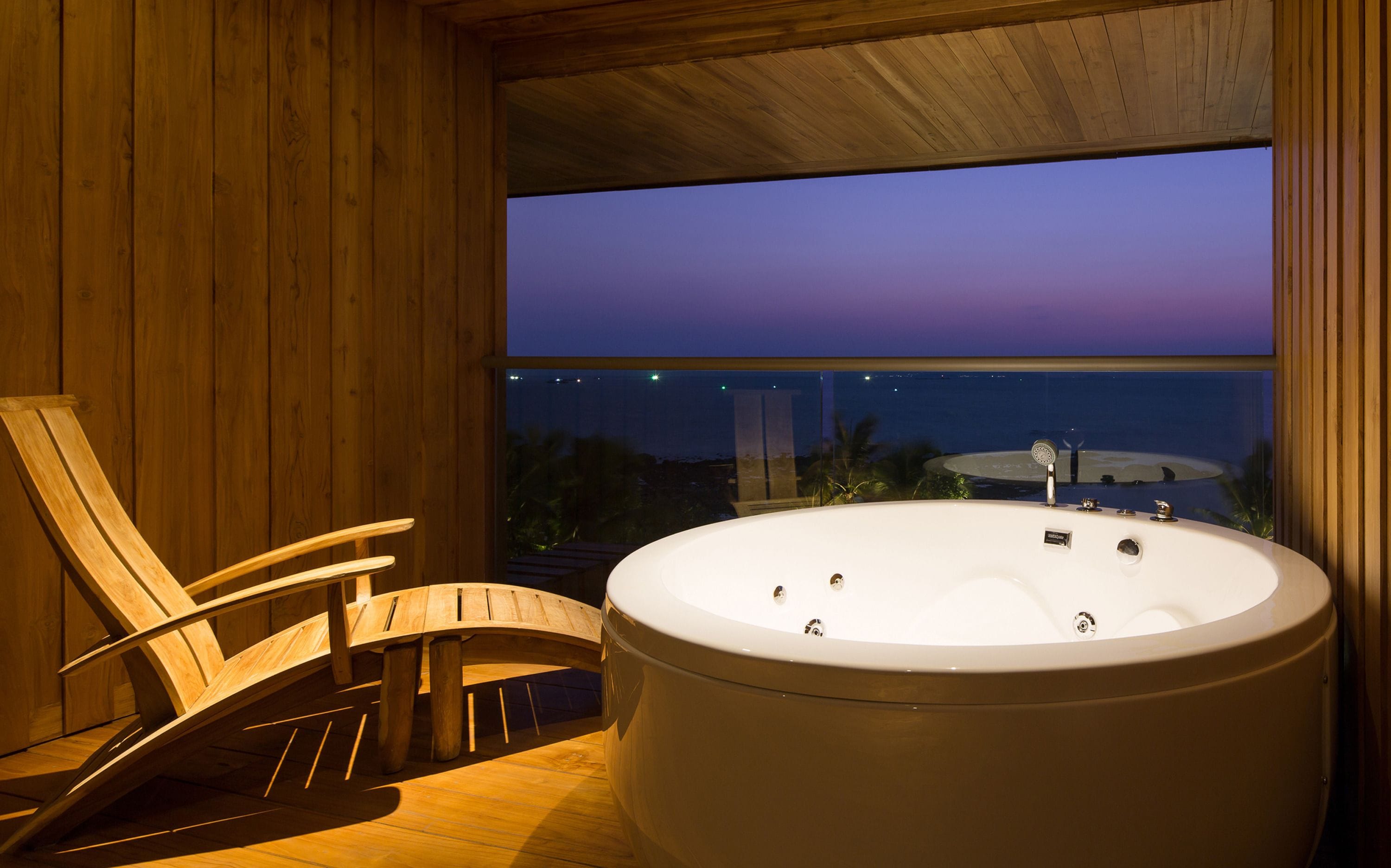 A wooden interior with a large bathtub and a wooden chair by a window overlooking a beach at dusk.