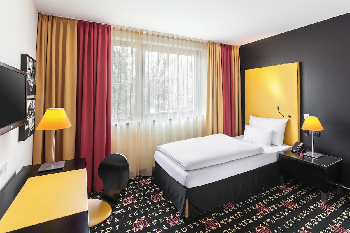 A hotel room with a bed, bedside table, lamp, TV, and a desk with a chair.