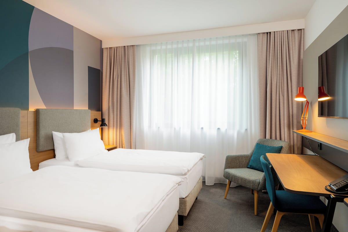 A hotel room with two beds, a desk, a chair, and a large window with curtains.