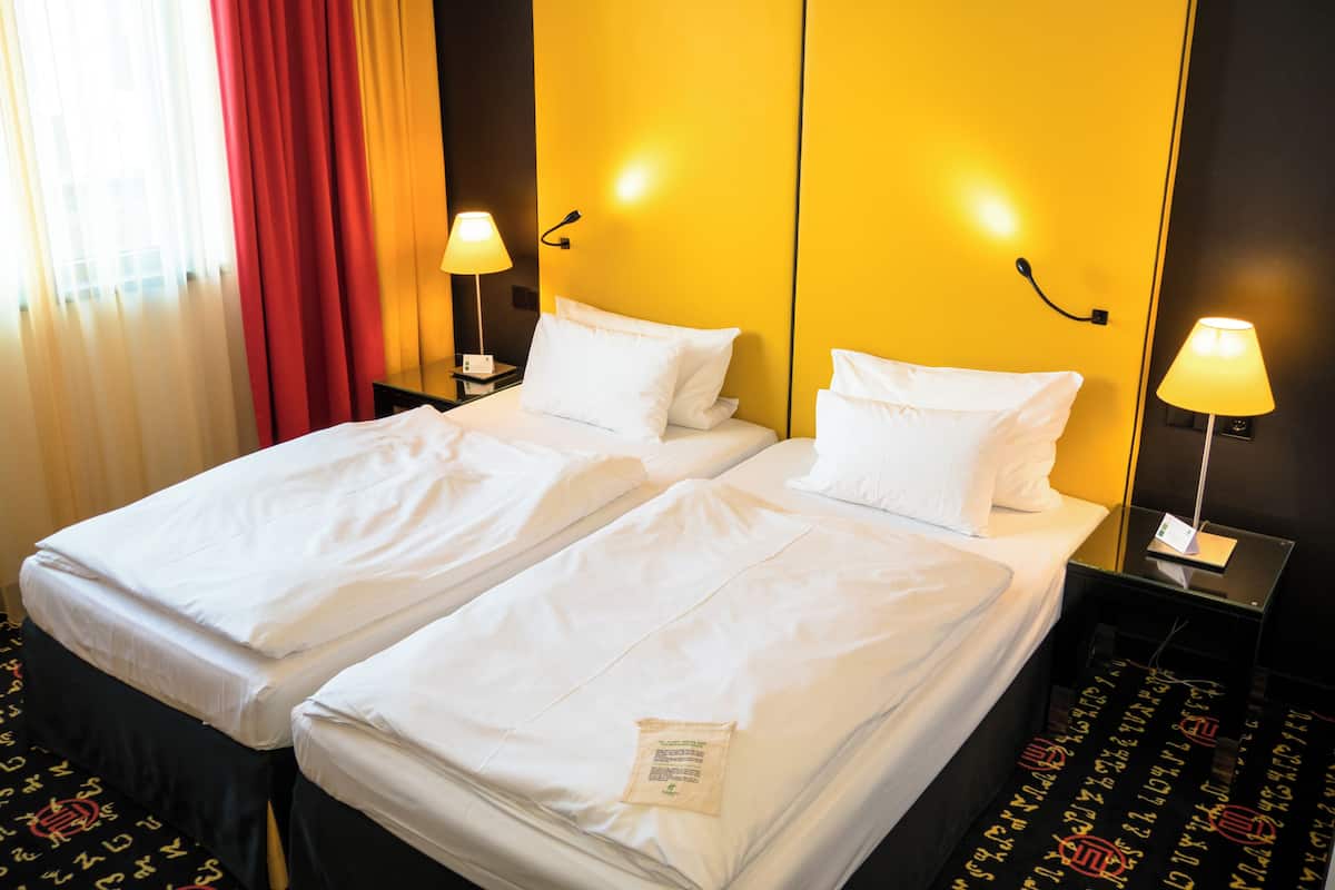 A hotel room with two single beds, a nightstand with a lamp, and a red curtain.