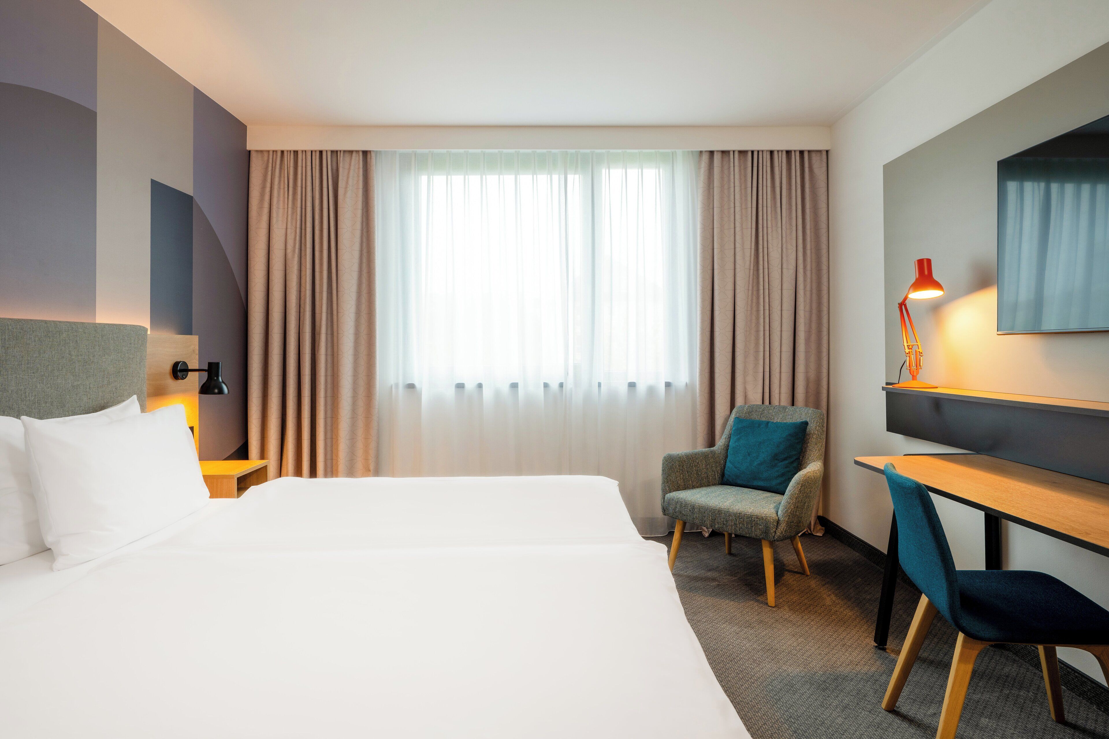 A hotel room with a large bed, a desk, a chair, and a lamp.