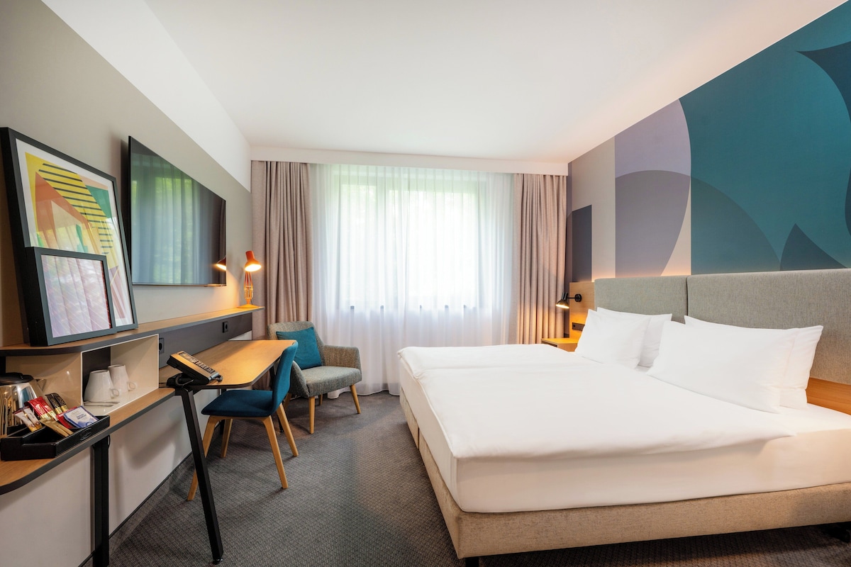 Una camera d'albergo con un letto grande, una scrivania con sedia, una televisione e una finestra con tende.