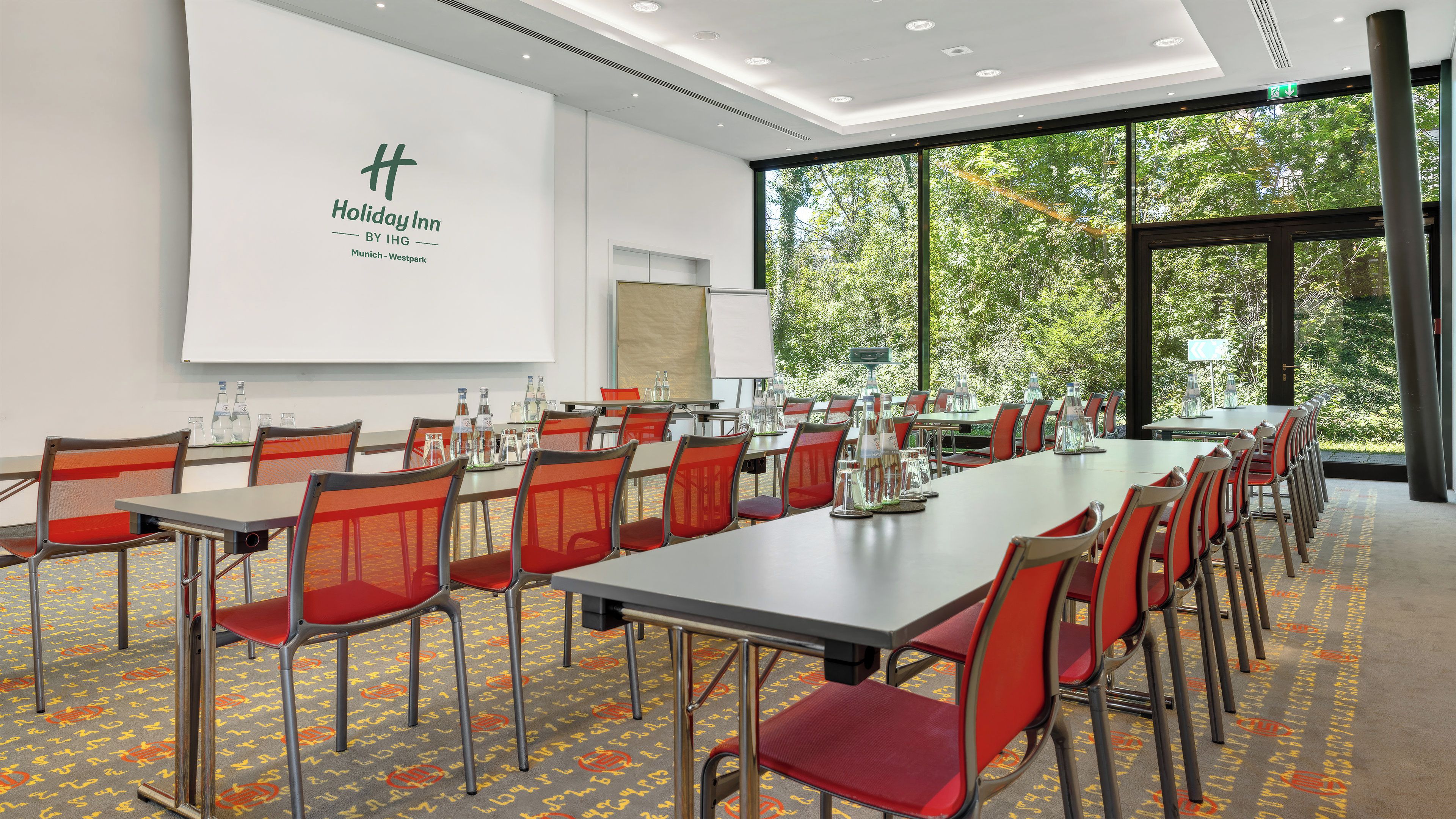 Un salón de conferencias con una mesa larga, sillas rojas y una pantalla de proyector.