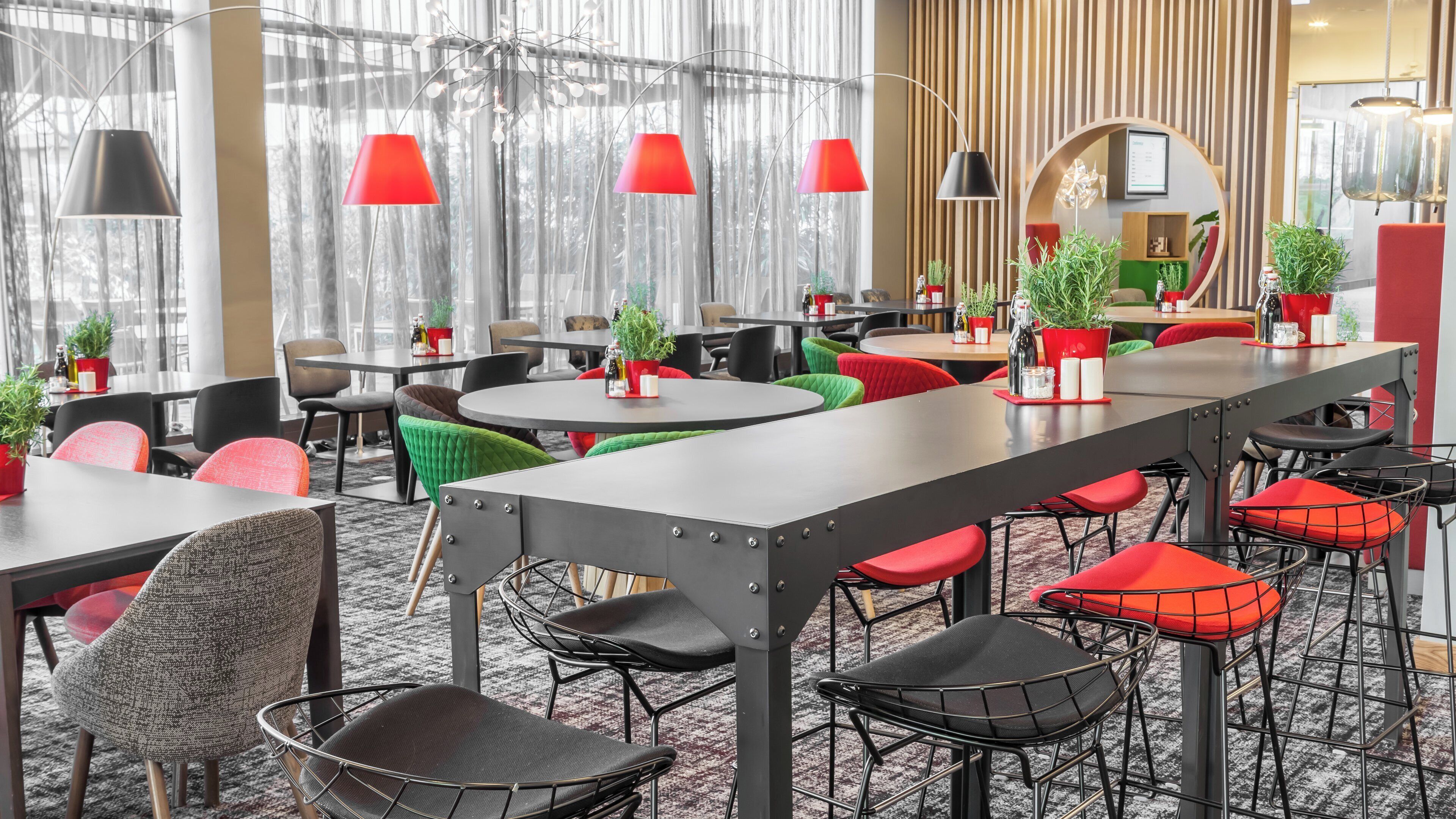 A modern hotel dining area with round tables, red and black chairs, and potted plants.