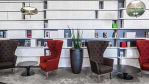 A modern library with bookshelves, red and brown upholstered chairs, and a central potted plant.