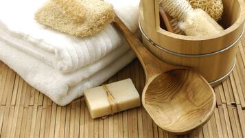 A wooden tub with a sponge and a wooden scoop, a bar of soap, and neatly folded white towels on a wooden floor.
