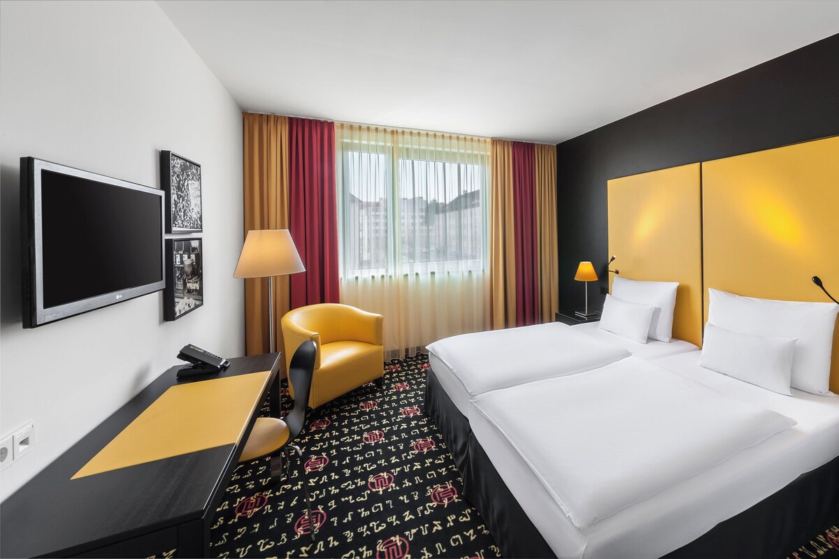Habitación de hotel con cama, dos mesitas de noche, escritorio, silla, televisor y ventana con cortinas.