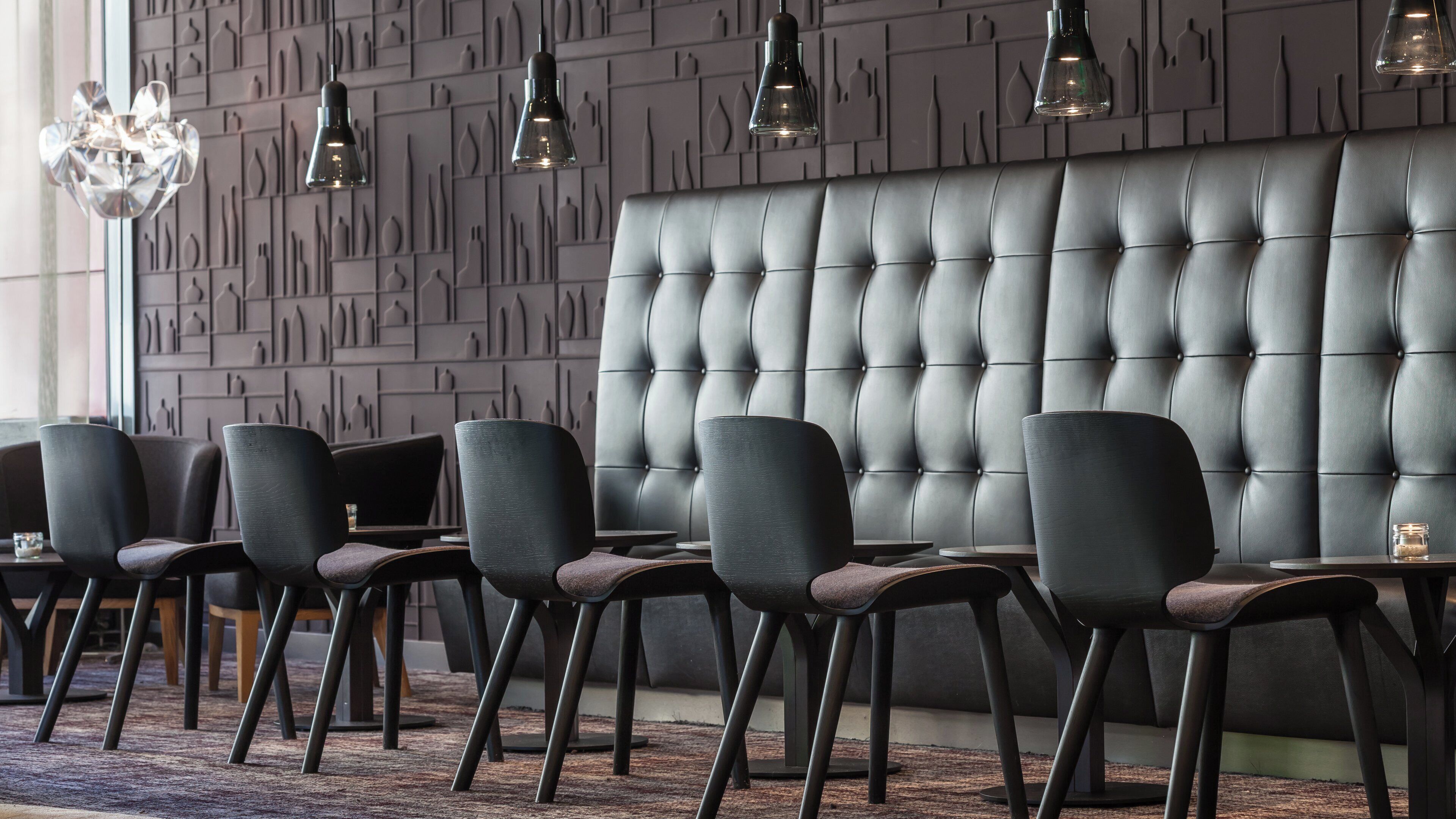 A modern restaurant interior with a tufted leather booth, dark chairs, and pendant lighting.