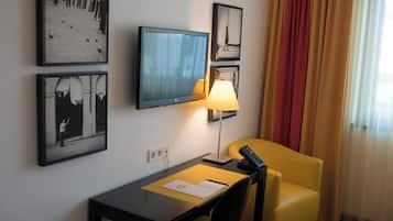 A hotel room with a desk, chair, TV, lamp, and framed pictures on the wall.