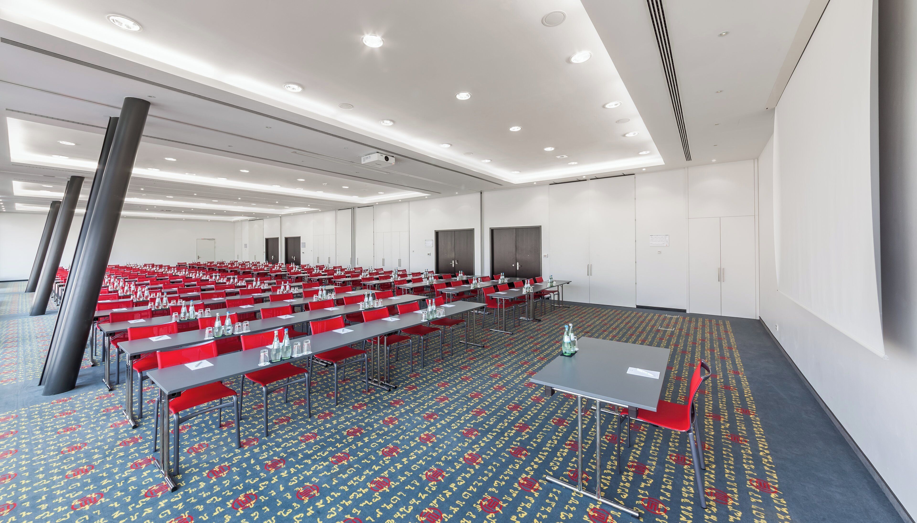 A conference room with rows of tables and chairs, a patterned carpet, and a white ceiling with recessed lighting.