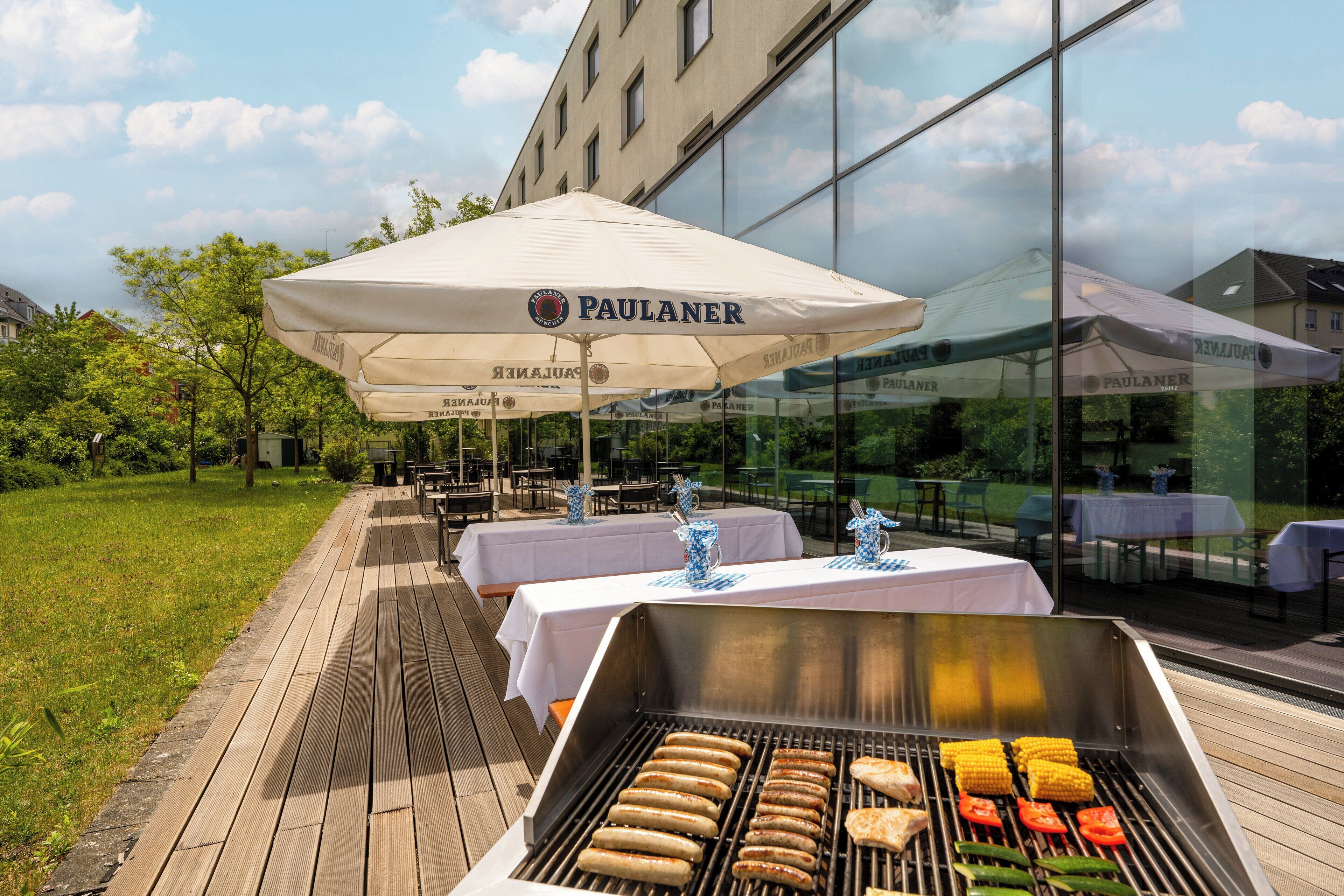 An outdoor dining area with tables and chairs, a grill with food, and umbrellas with the brand "PAULANER".