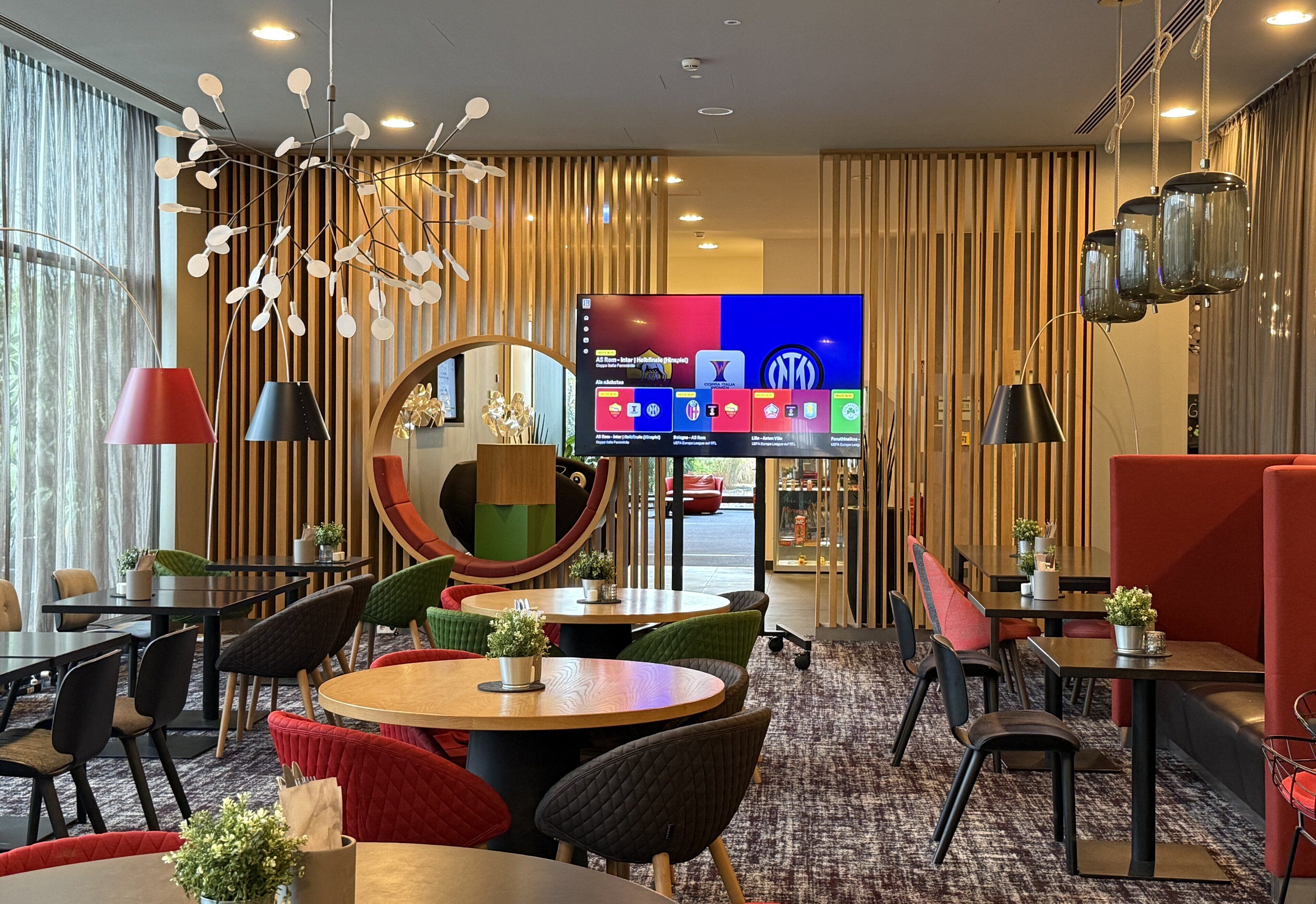 A modern hotel lobby with round tables, red and black chairs, and a large TV screen displaying hotel information.