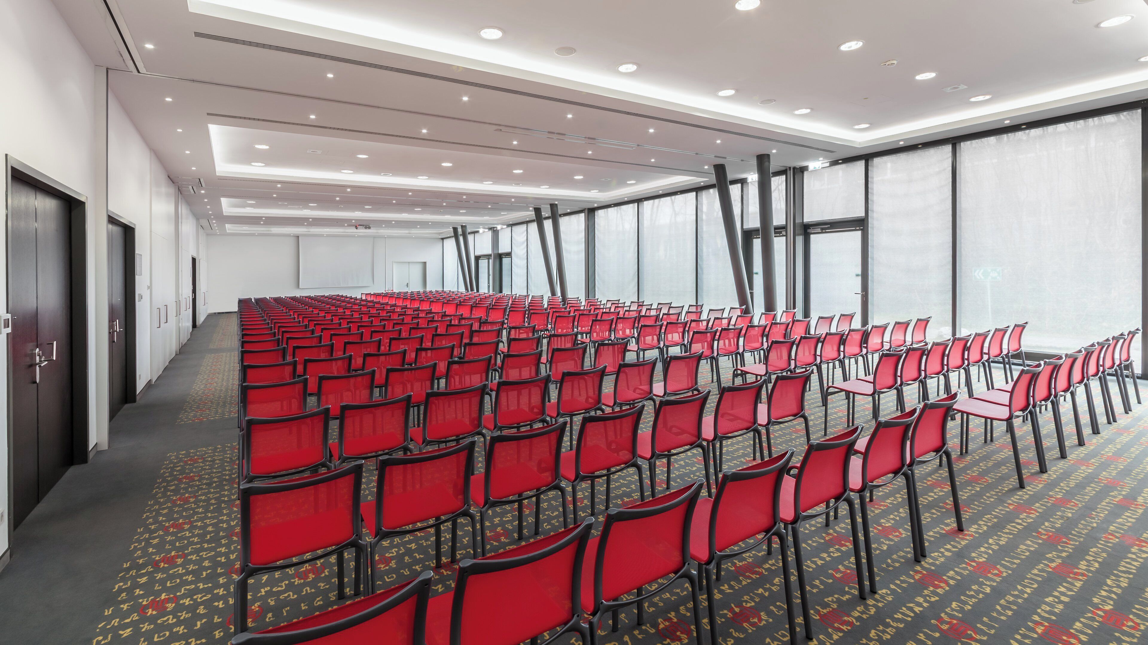 A conference room with rows of red chairs, a patterned carpet, and large windows with curtains.