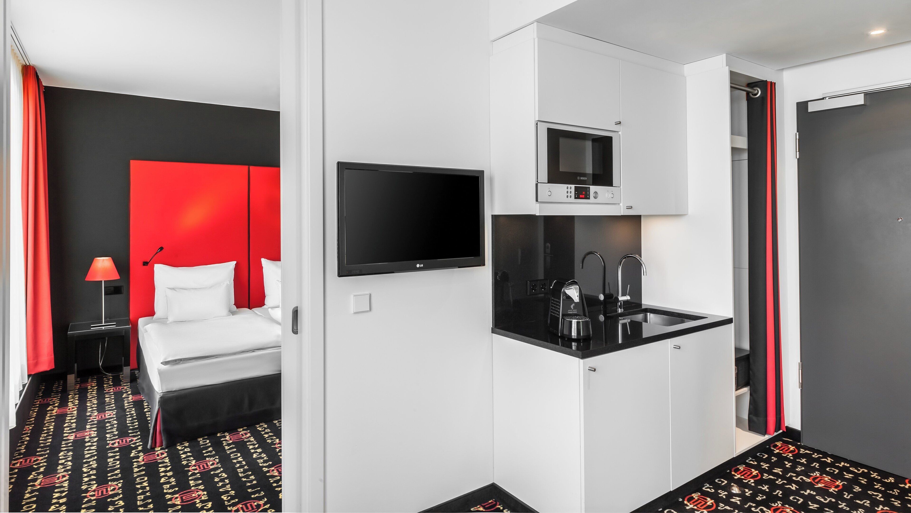A hotel room with a bed, a TV, a kitchenette, and a red accent wall.