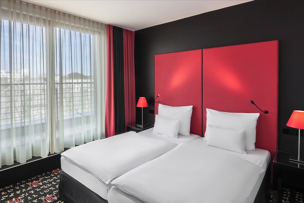 Una camera d'albergo con un letto grande, due comodini, lampade rosse e tende leggere.