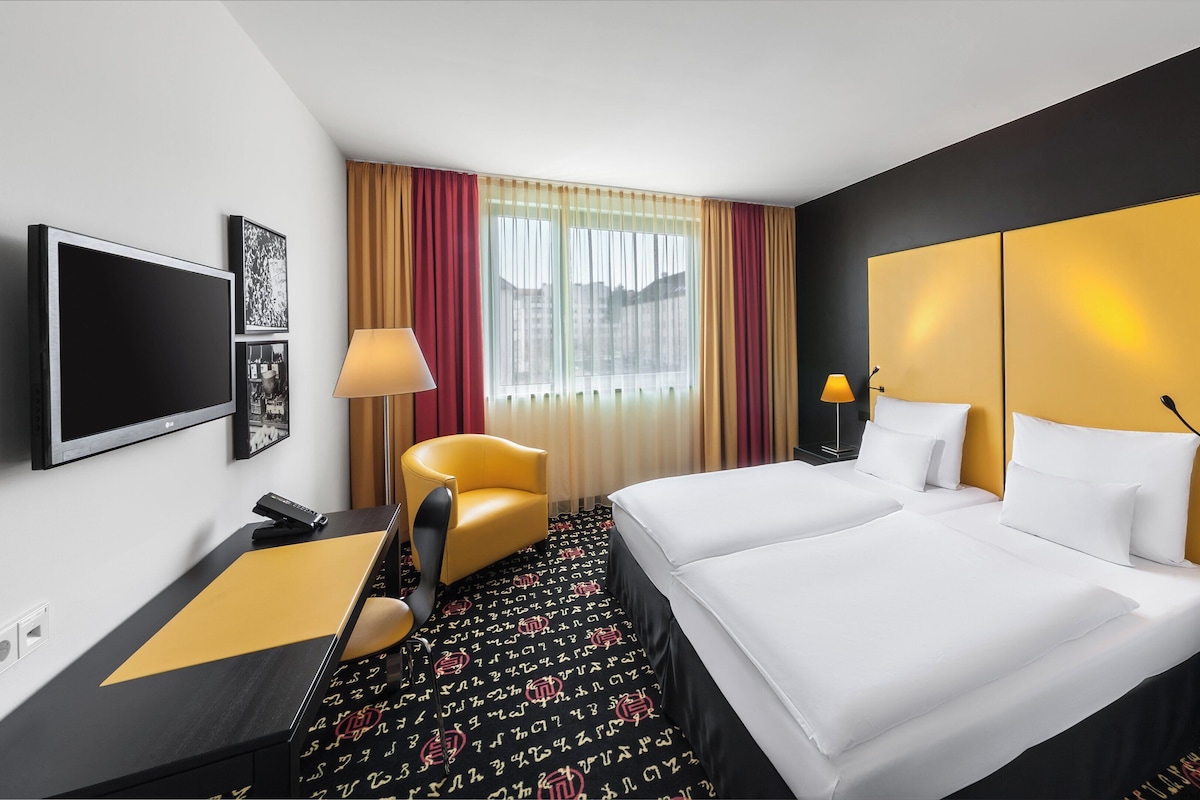 Habitación de hotel con cama, dos mesitas de noche, escritorio, silla, televisor y ventana con cortinas.
