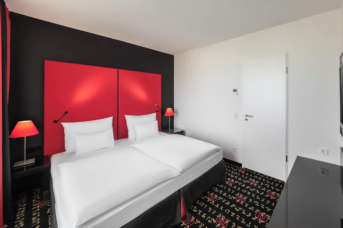 A hotel room with a red headboard, white bedding, a flat-screen TV, and a door with a handle.