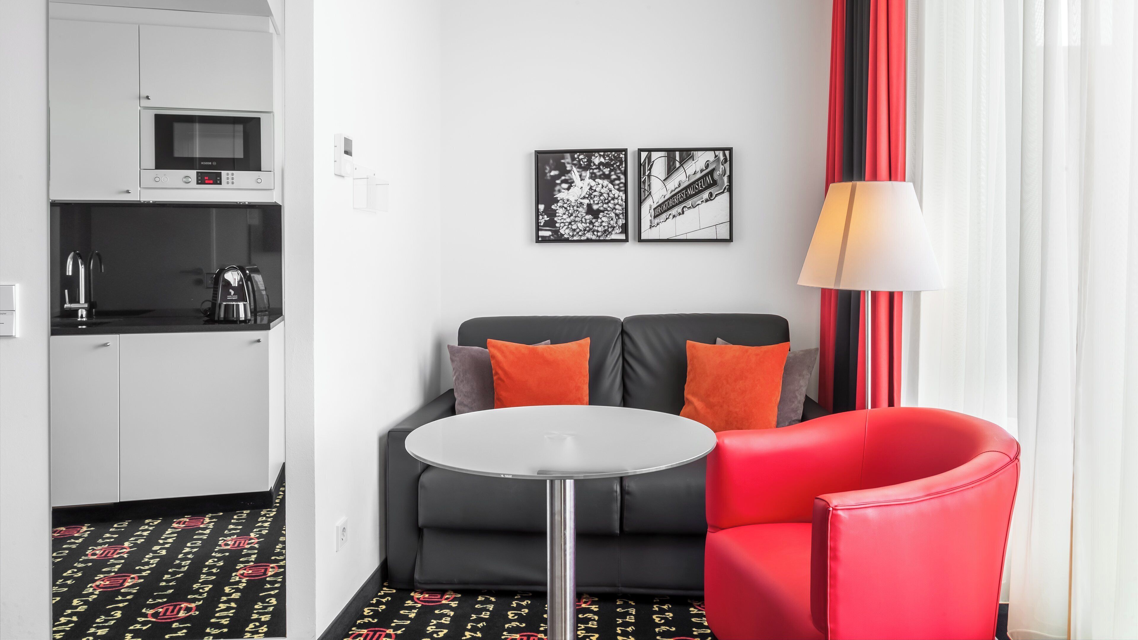 A modern hotel room with a kitchenette, a sofa with orange cushions, a red armchair, and two framed pictures on the wall.