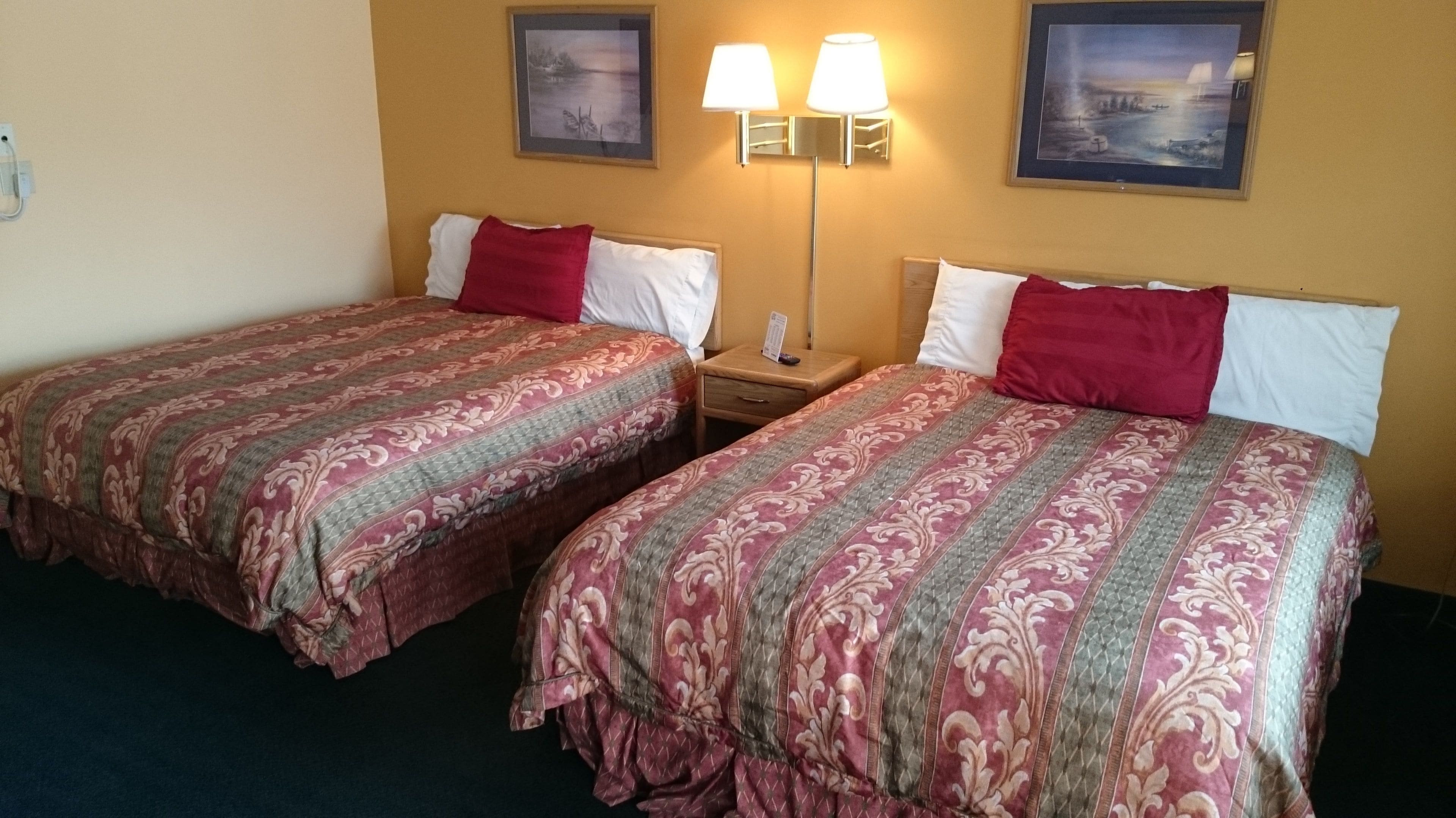 A hotel room with two beds, each with a patterned bedspread and red pillows. There are two wall-mounted lamps and two framed pictures on the wall.